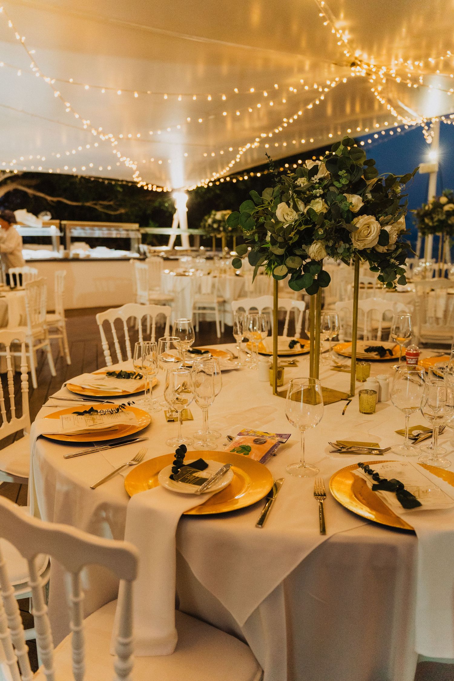 Elegant wedding reception table with gold charger plates, white linens and twinkle lights strung overhead at an outdoor venue.