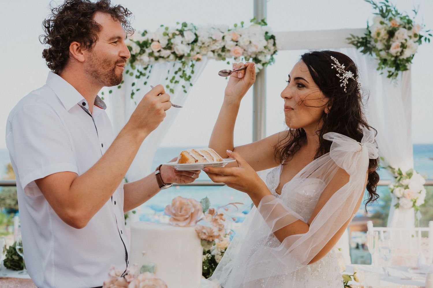 Newlyweds playfully feed each other wedding cake at their beach reception.