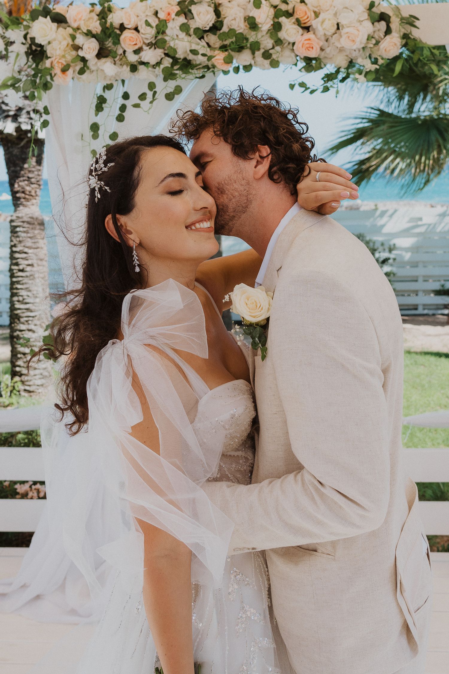 Tender moment between newlyweds under floral wedding arch with seaside backdrop.