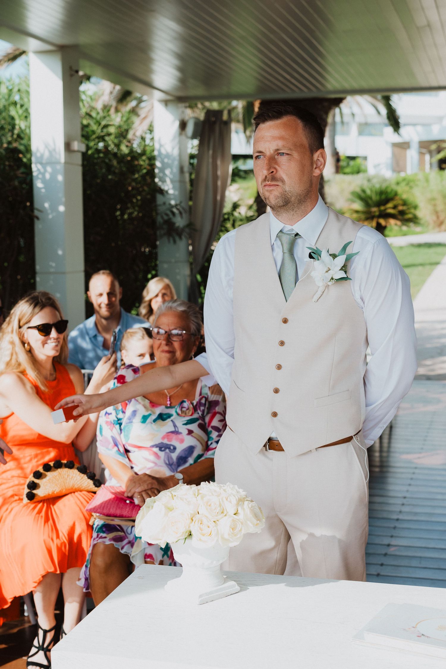 A groom in a light beige vest and tie waits at a covered outdoor venue with a white cake on the table.