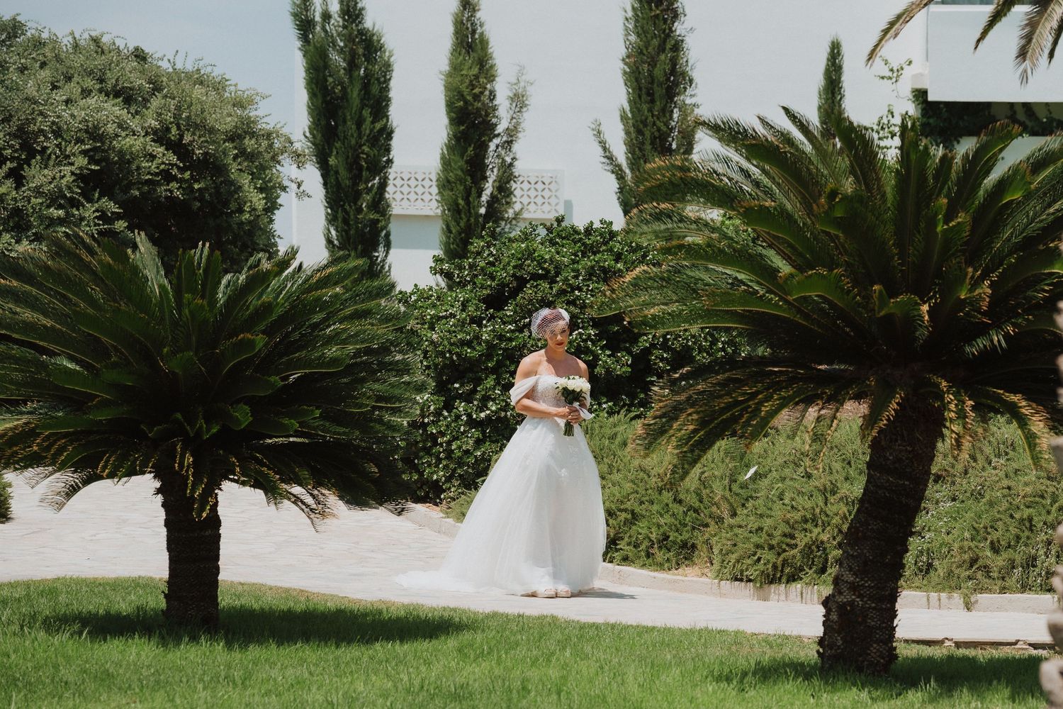 A white wedding dress flows between palm trees on a manicured lawn with ocean views.