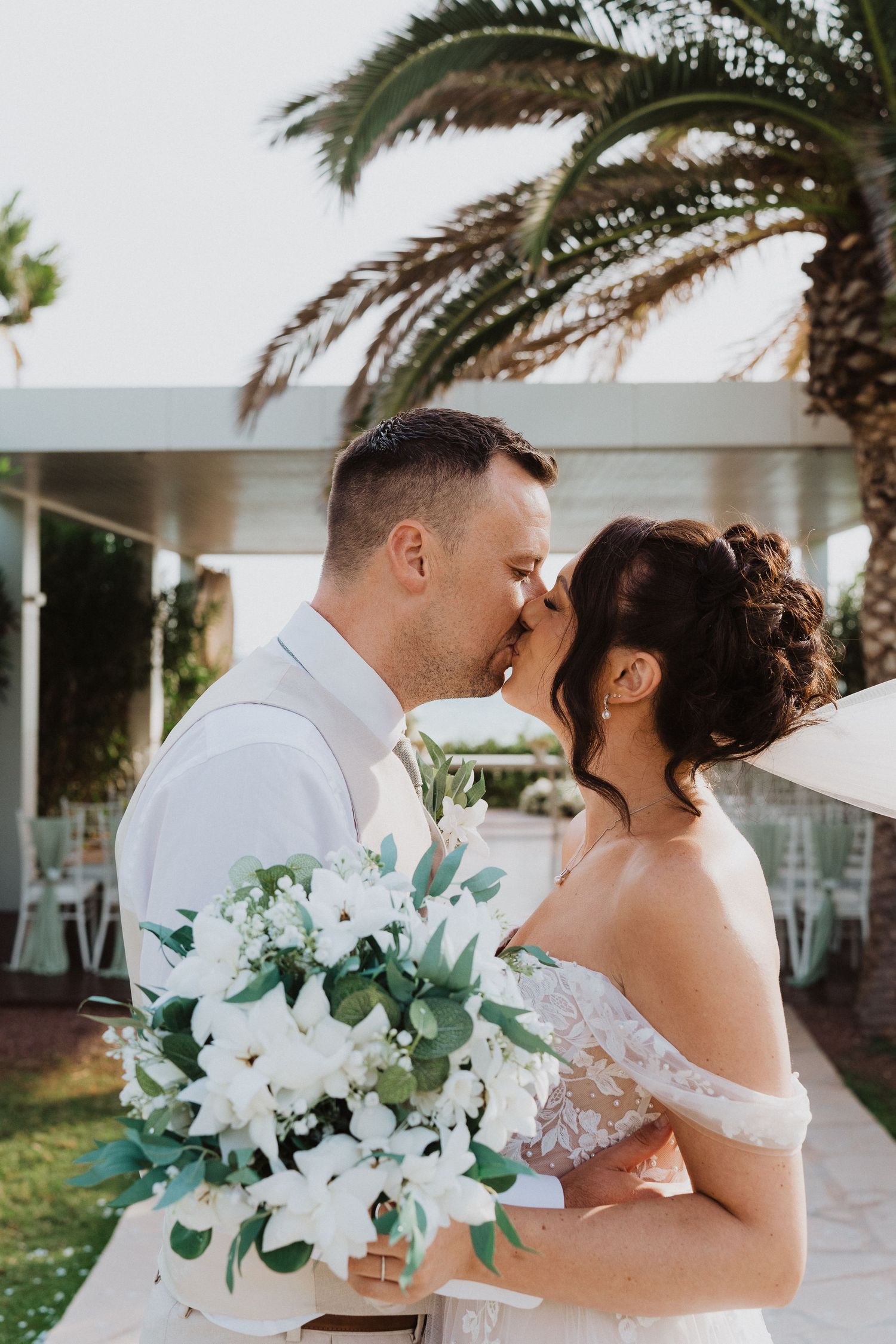 A romantic wedding moment outdoors with palm trees, featuring a white floral bouquet and off-shoulder lace dress.