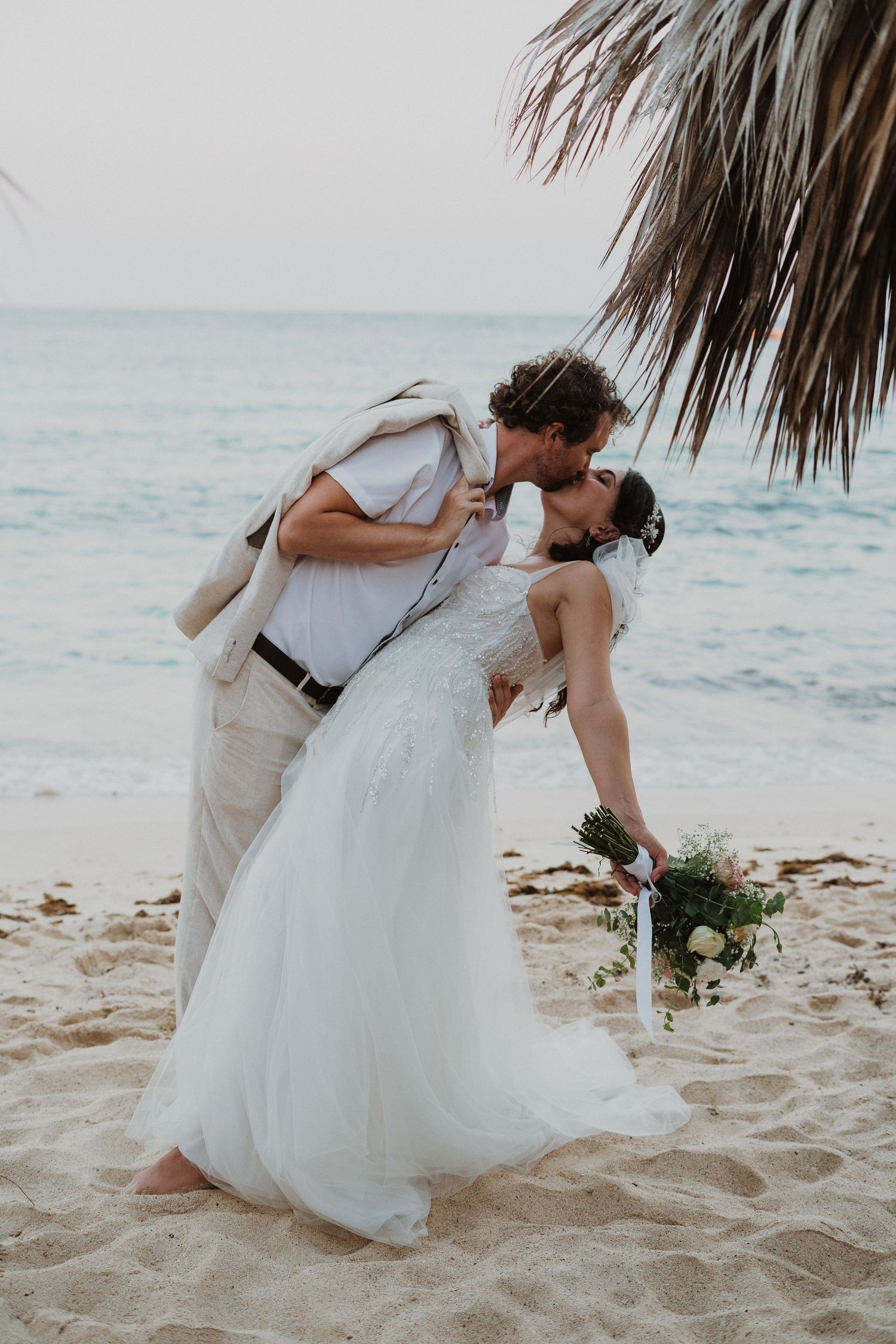 Dramatic beach wedding kiss pose with turquoise ocean and palm leaves in background.