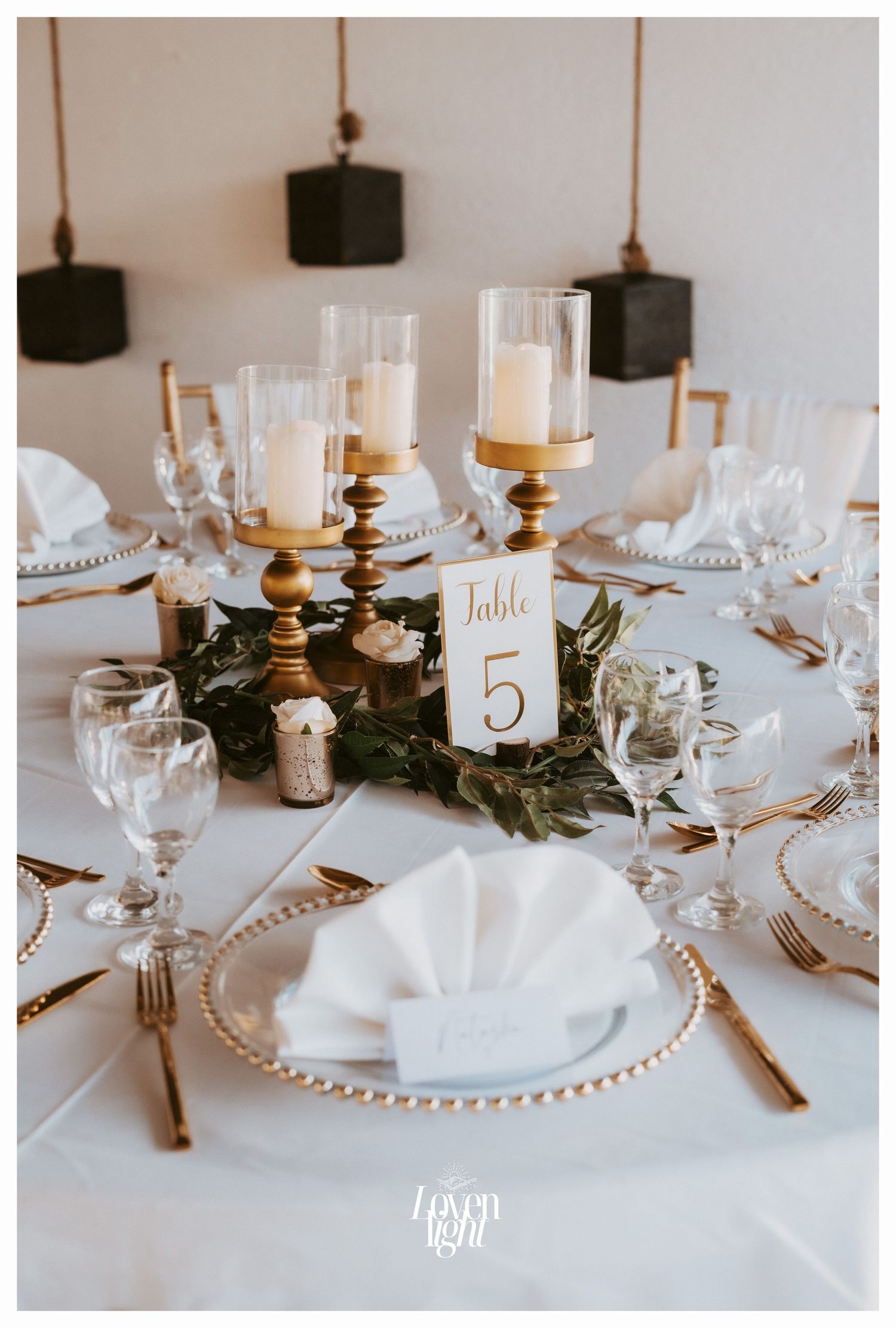Wedding table centerpiece with gold candle holders, greenery and white place settings.