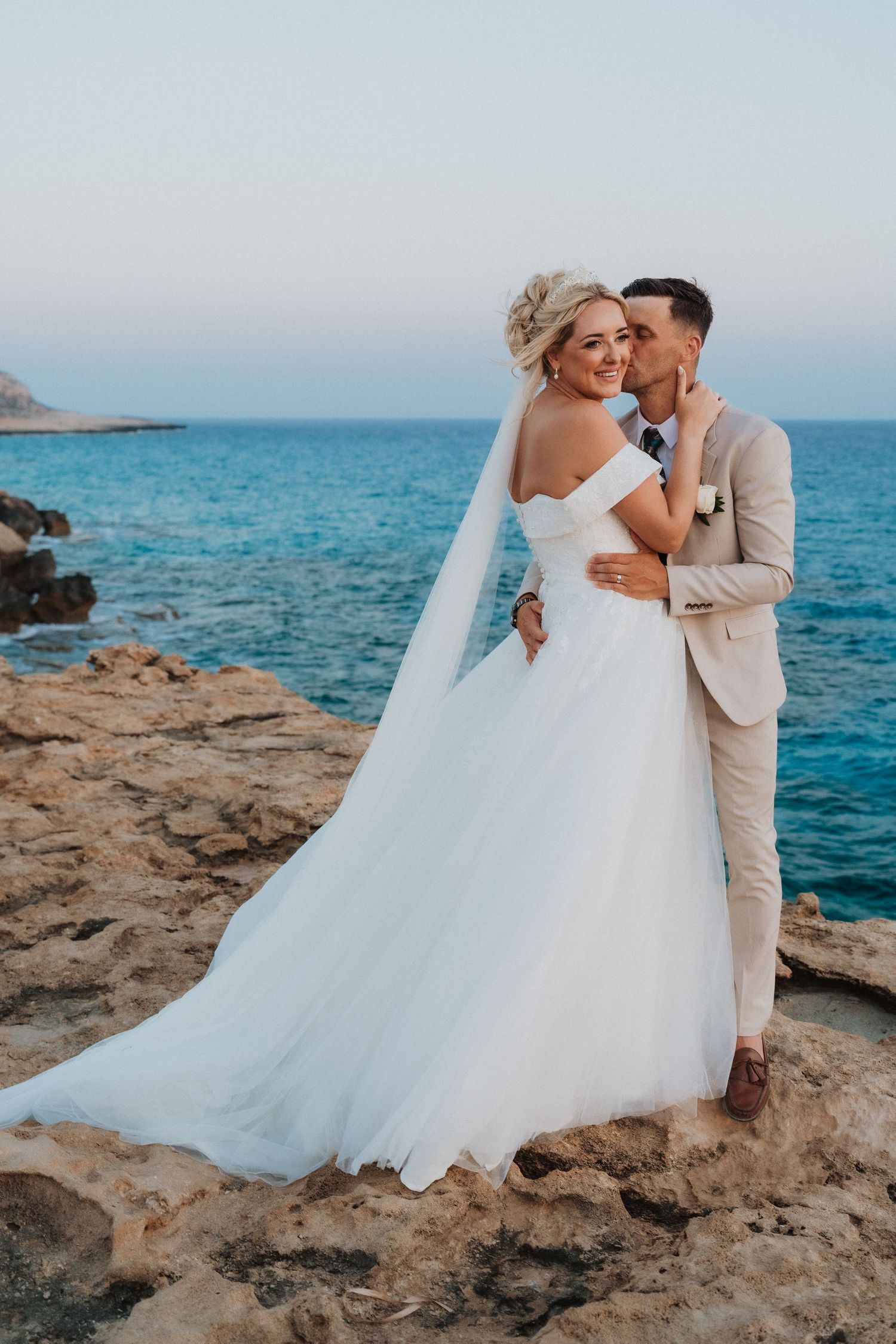 A romantic coastal wedding photo on rocky cliffs overlooking bright blue Mediterranean waters.