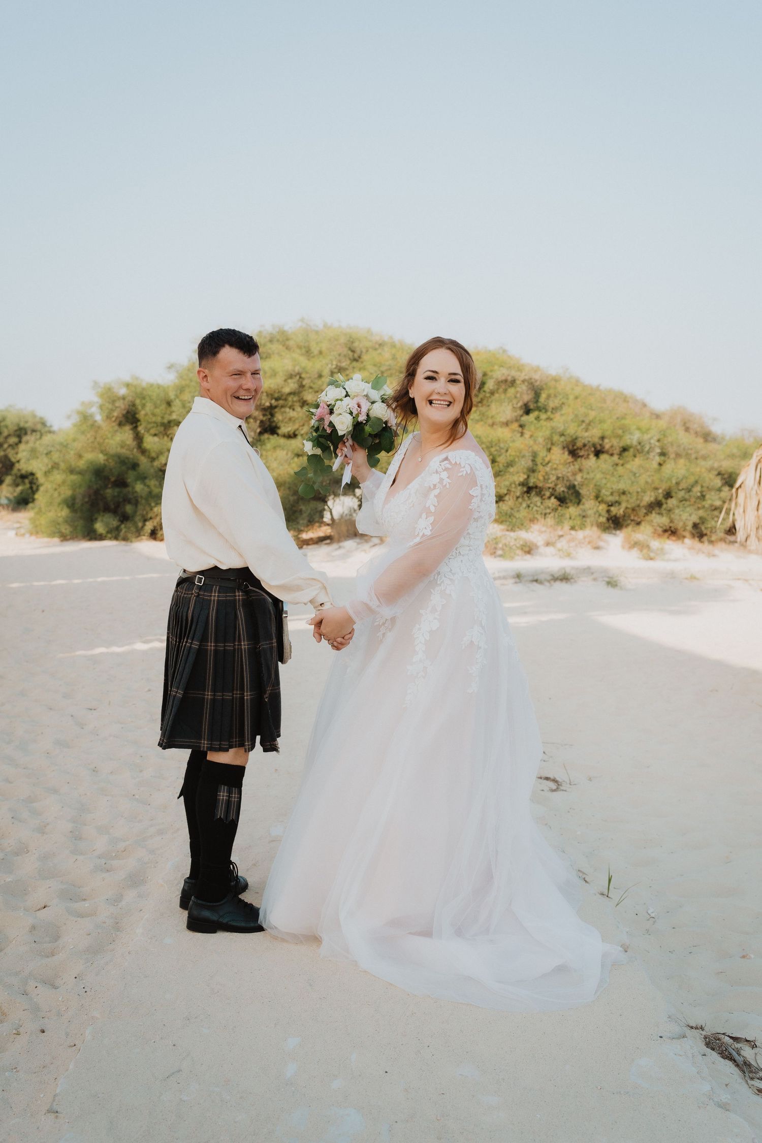 A couple poses on a beach, with the groom wearing a traditional Scottish kilt and bride in a flowing white dress.