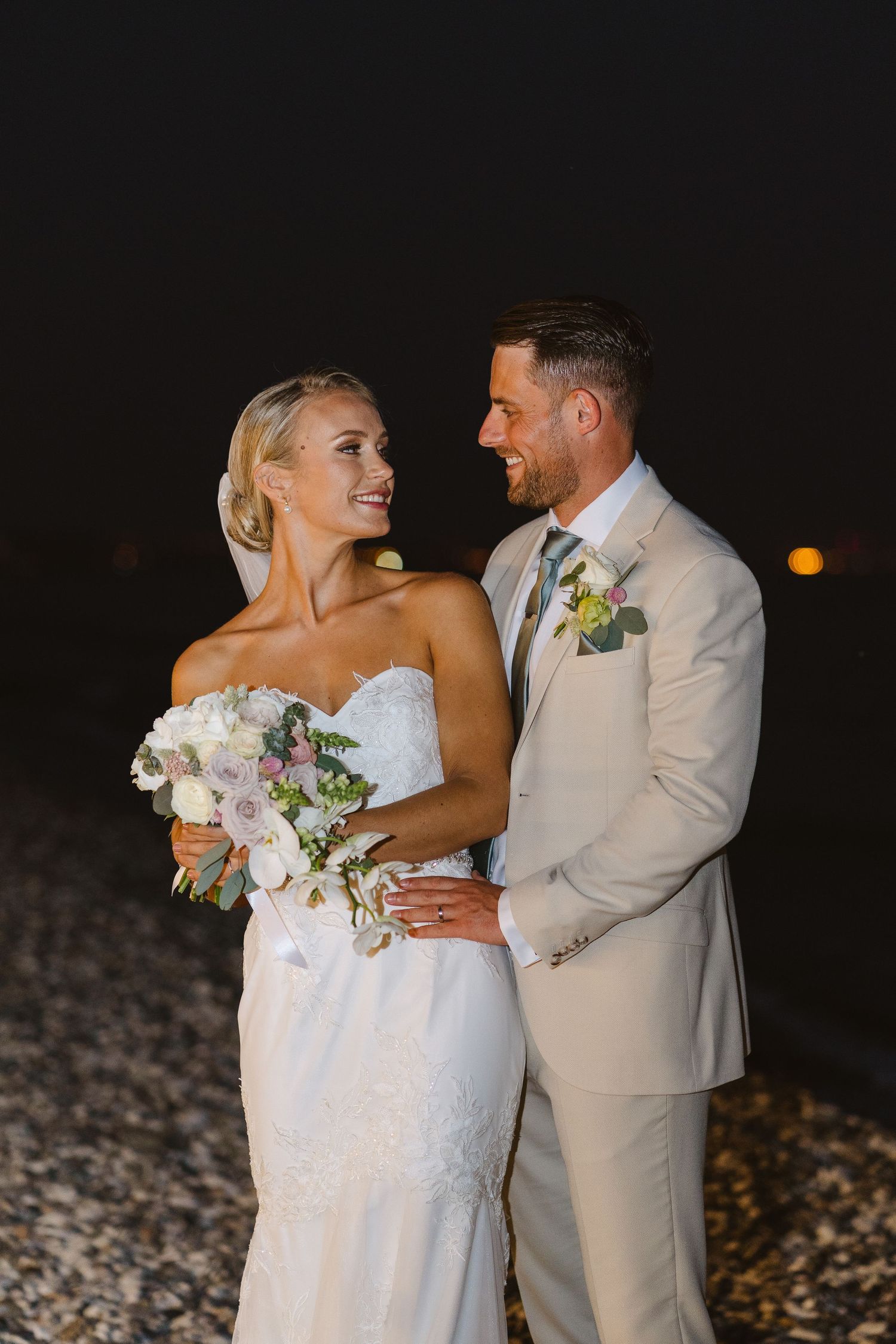 Romantic beach wedding photograph with couple in light formal wear against dark evening sky.