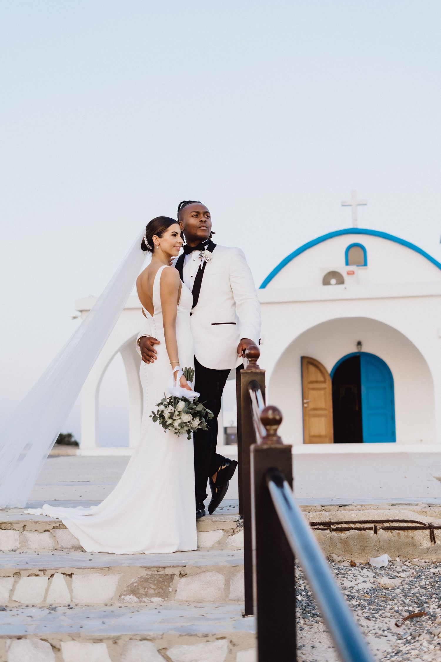 Elegant wedding couple poses on stone steps against white Mediterranean chapel architecture.