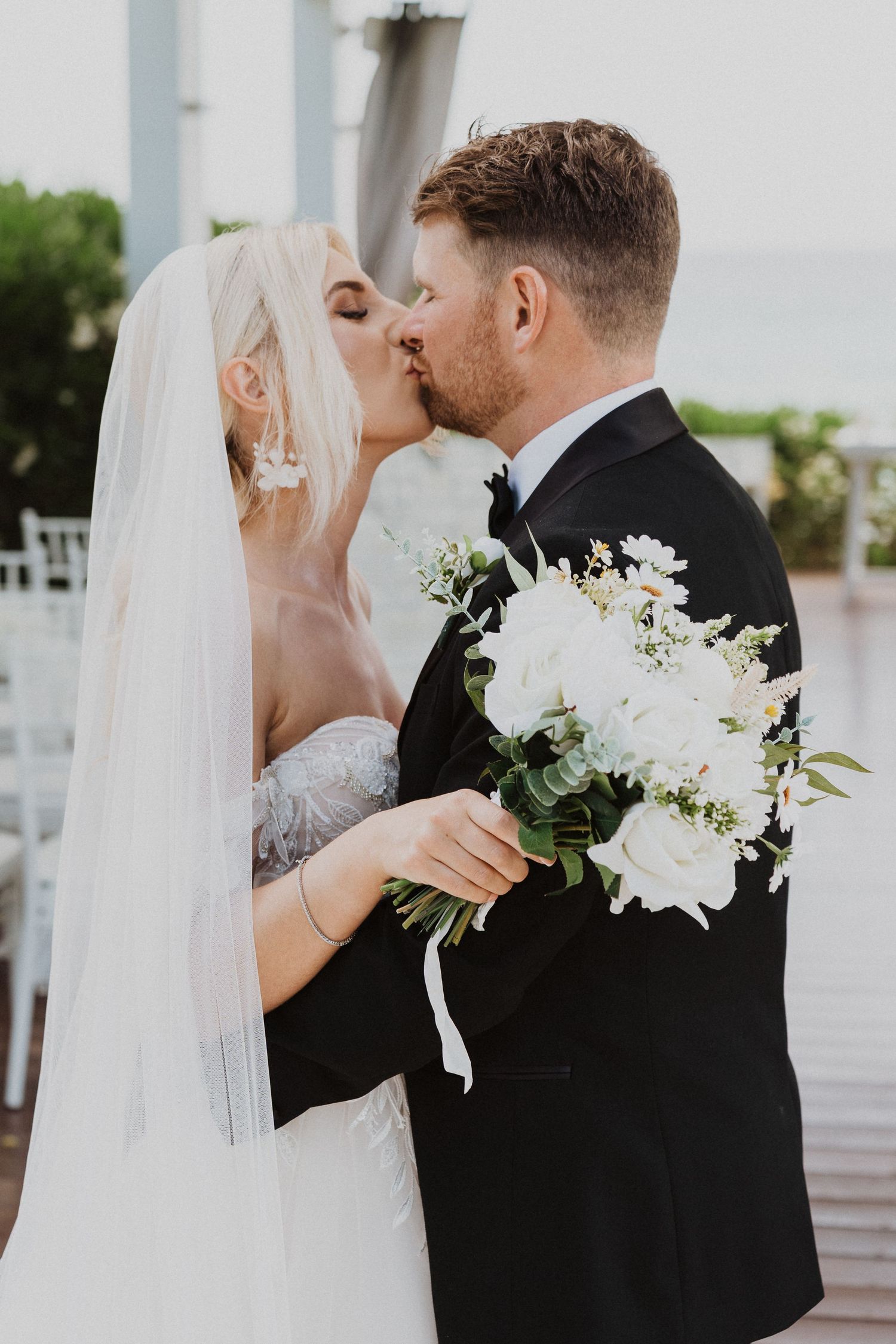 Newlyweds share a kiss while holding an elegant white floral bouquet at a seaside wedding ceremony.