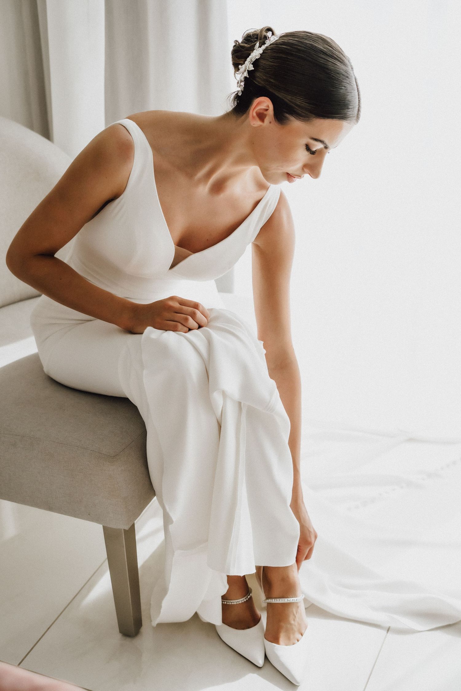 Elegant bride in white dress adjusting shoes while seated on grey chair.