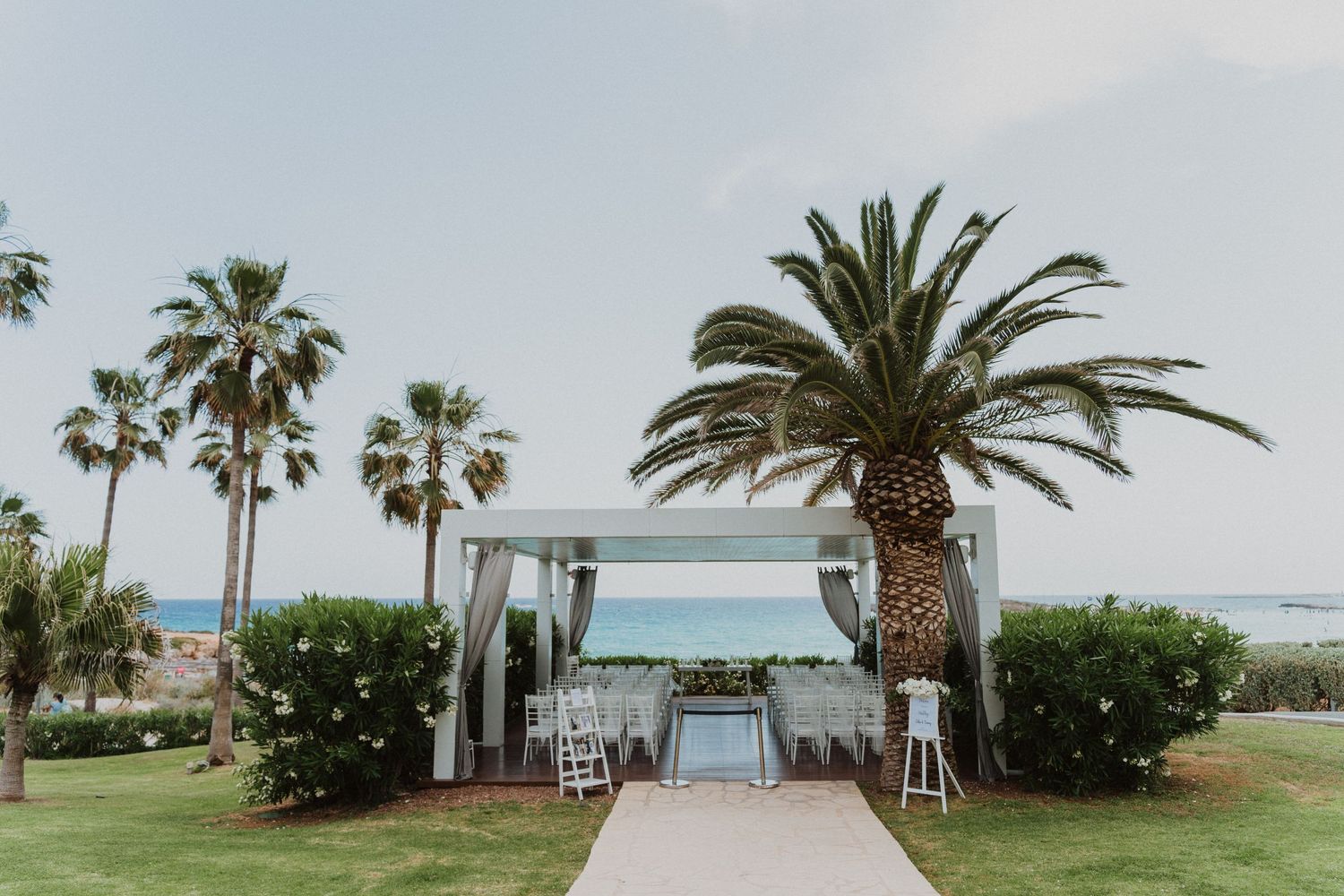 Beachfront wedding ceremony setup with palm trees and white chairs facing the ocean.
