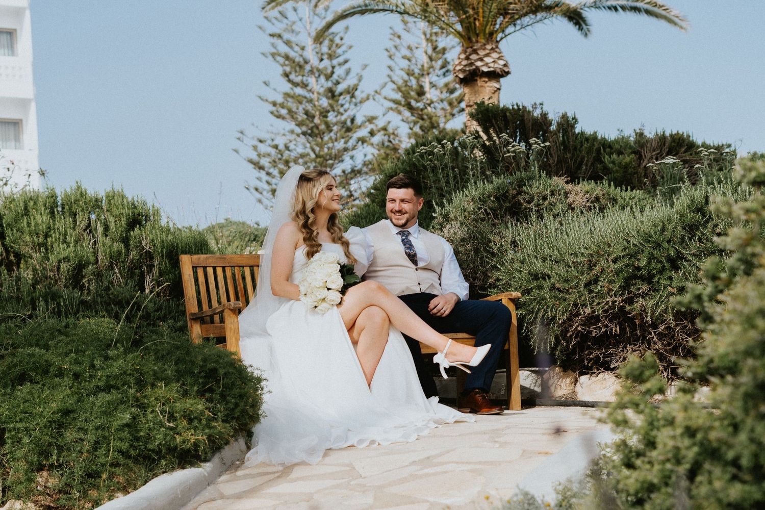 Couple sitting on wooden bench in lush garden setting with ocean view.