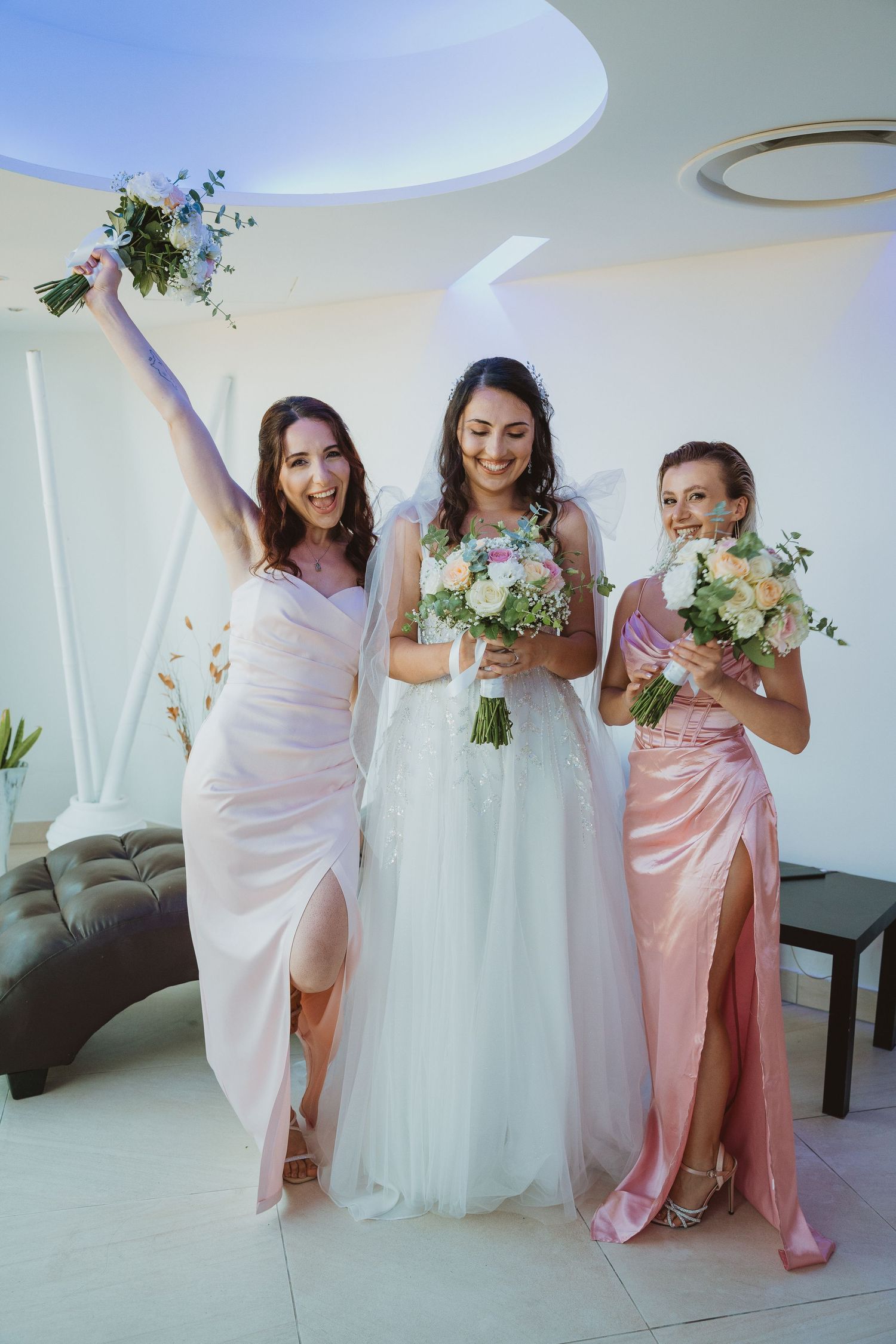 Bridesmaids in blush pink dresses stand with bride in white gown holding floral bouquets at indoor venue.