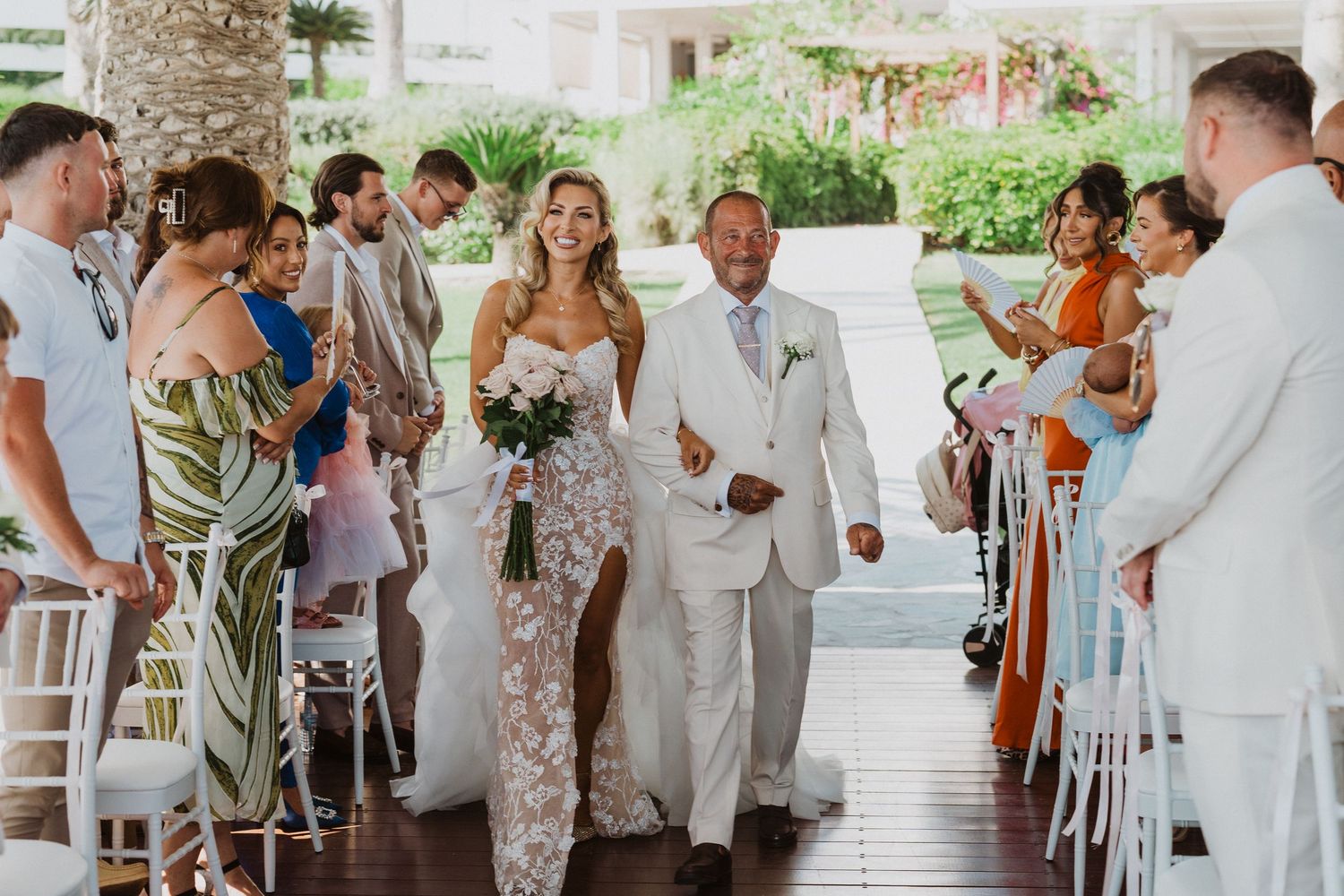 Wedding guests in light colored attire gather for a celebration at an outdoor venue with palm trees and white decor.