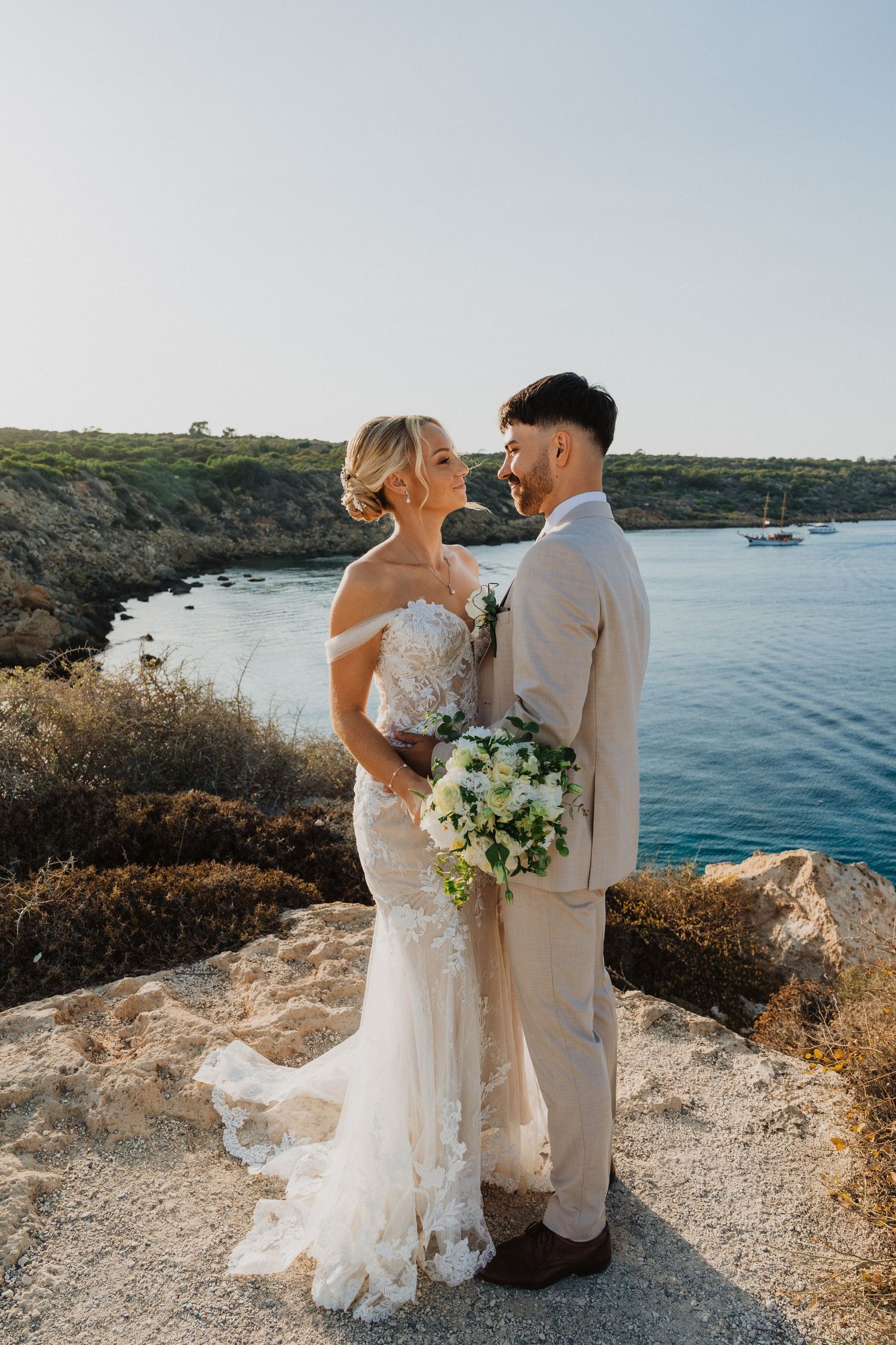 Elegant coastal wedding portrait with beige suit and lace dress against Mediterranean waters.