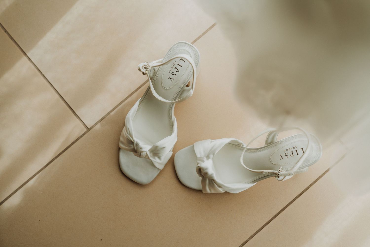 White wedding sandals with knotted straps photographed on beige floor.