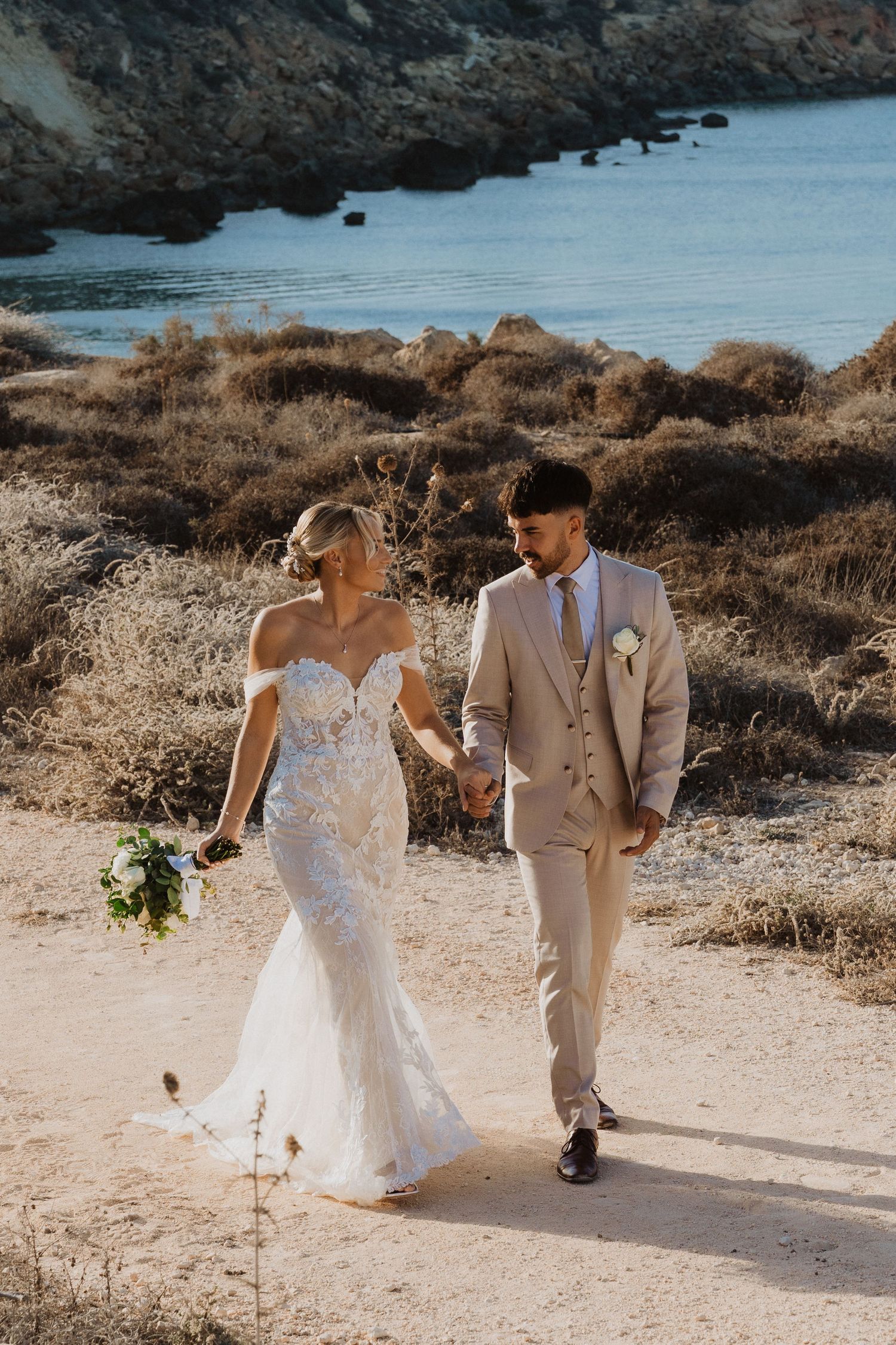 A wedding couple strolls hand in hand along a coastal pathway with dramatic seaside scenery.