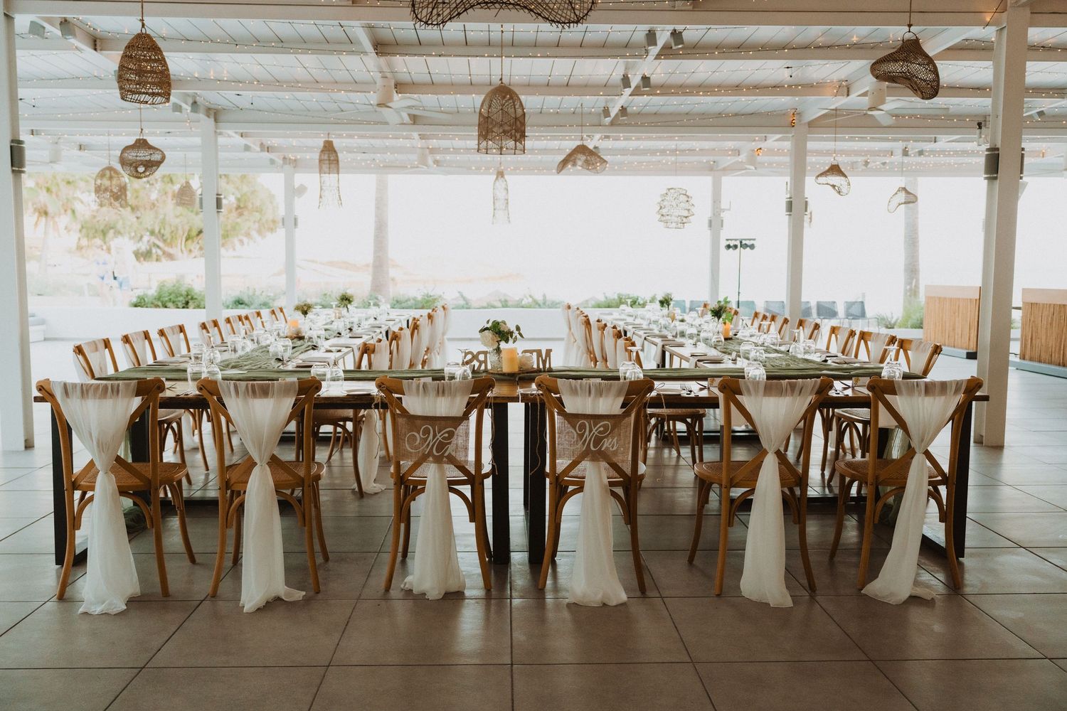 Long banquet tables set for a wedding reception under a covered outdoor pavilion with hanging lights.