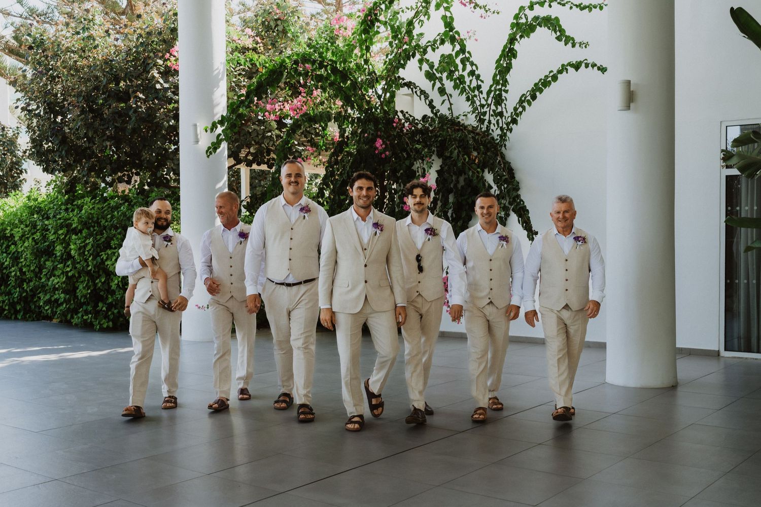 Wedding party in coordinated cream suits lined up against resort wall.
