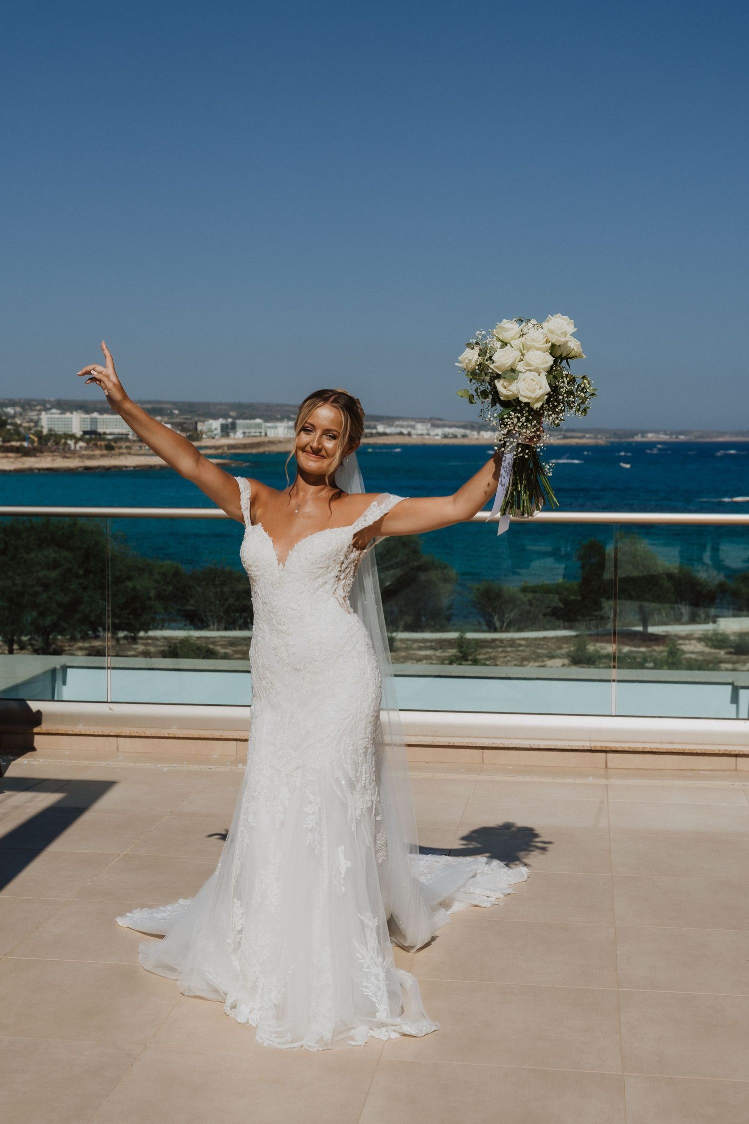 A bride in a white lace gown poses joyfully on a terrace overlooking a beautiful blue ocean coastline.