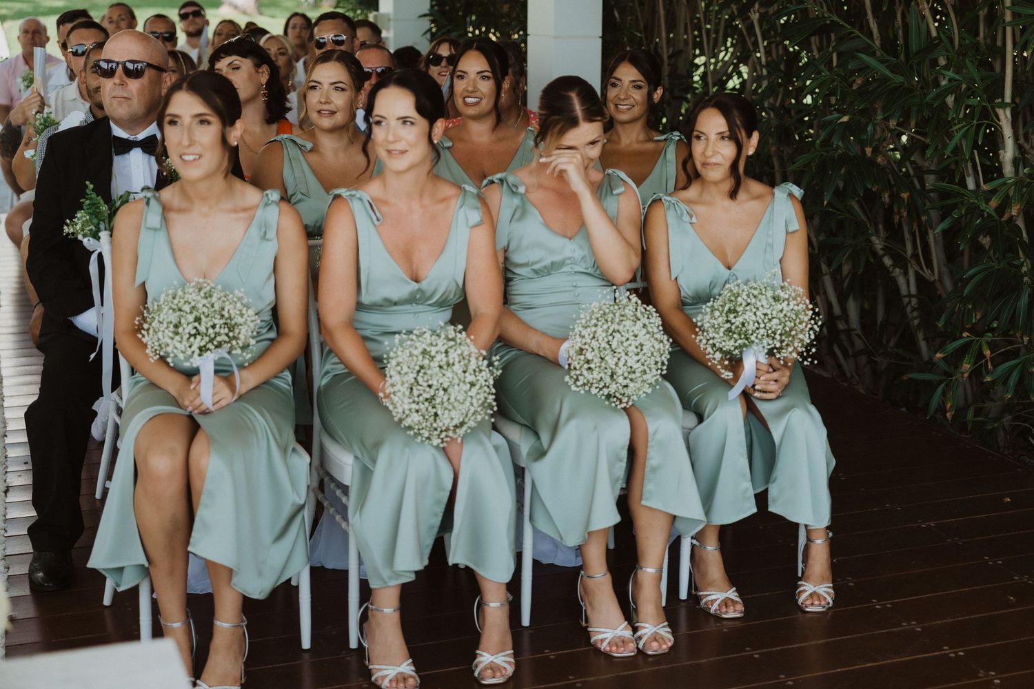 Bridesmaids in light blue dresses sit holding white bouquets during ceremony.