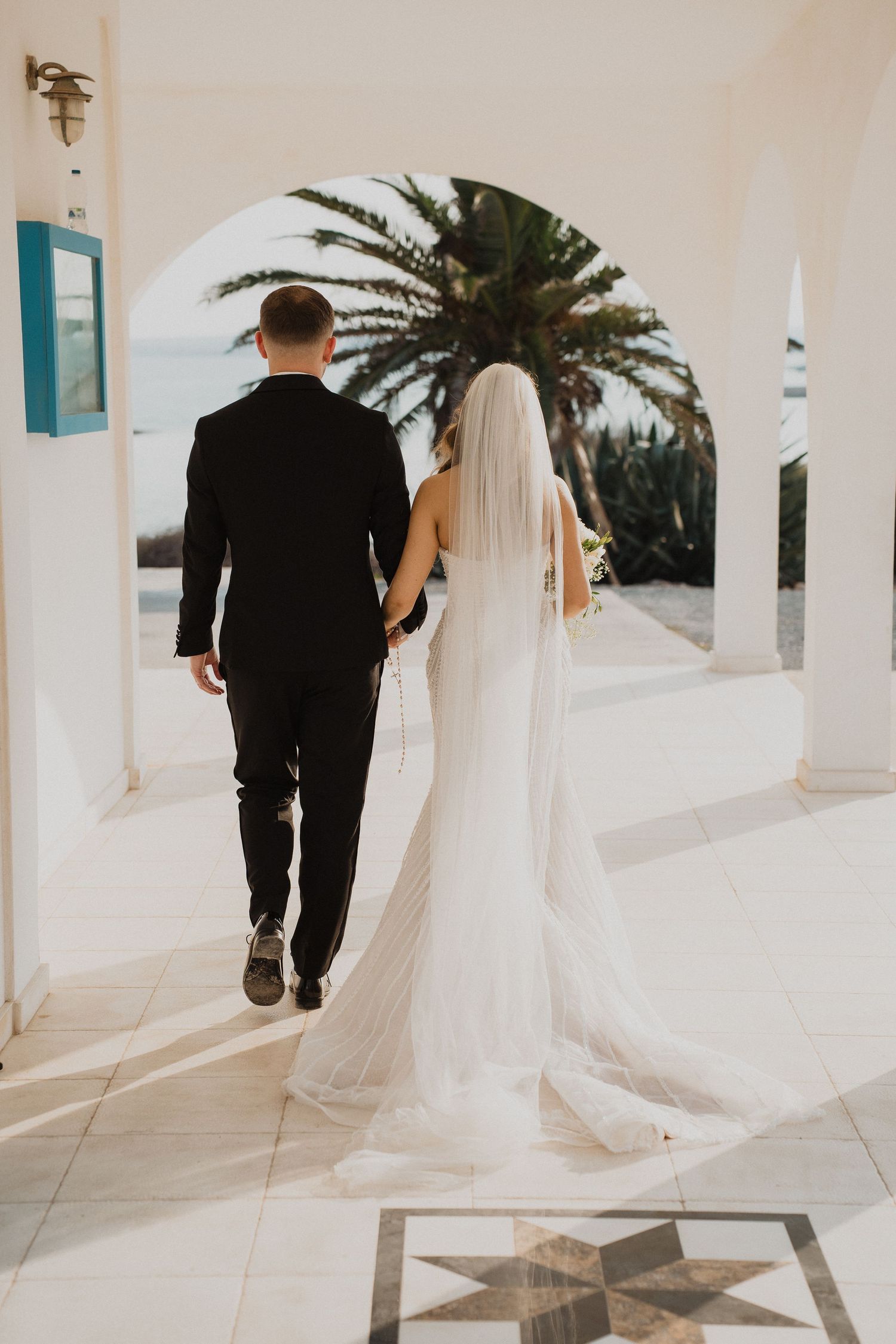 Bride and groom walk hand in hand through white arched corridor with ocean view and palm tree.