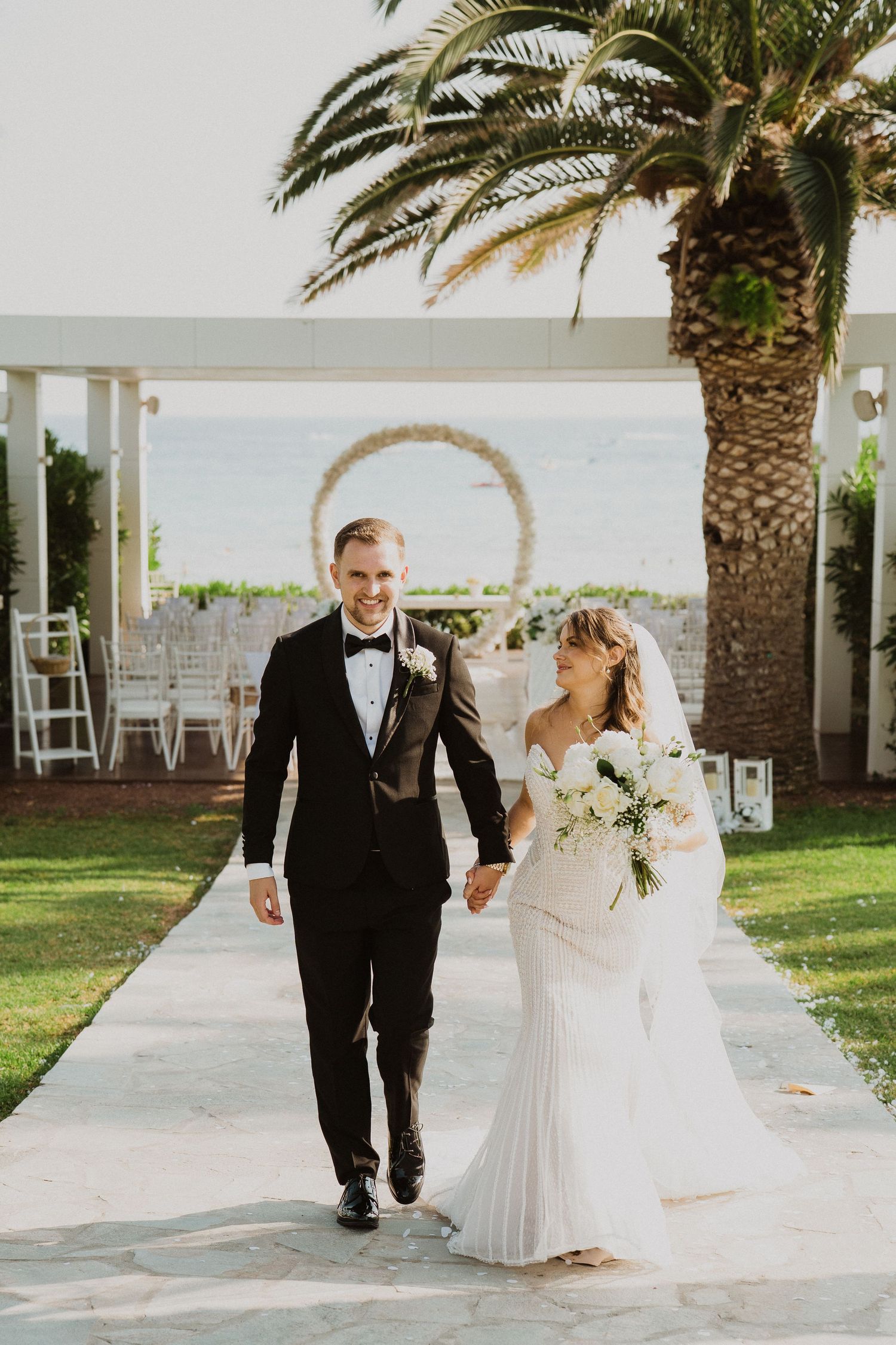 Smiling bride and groom walk hand in hand down aisle with circular arch and palm tree at seaside venue.