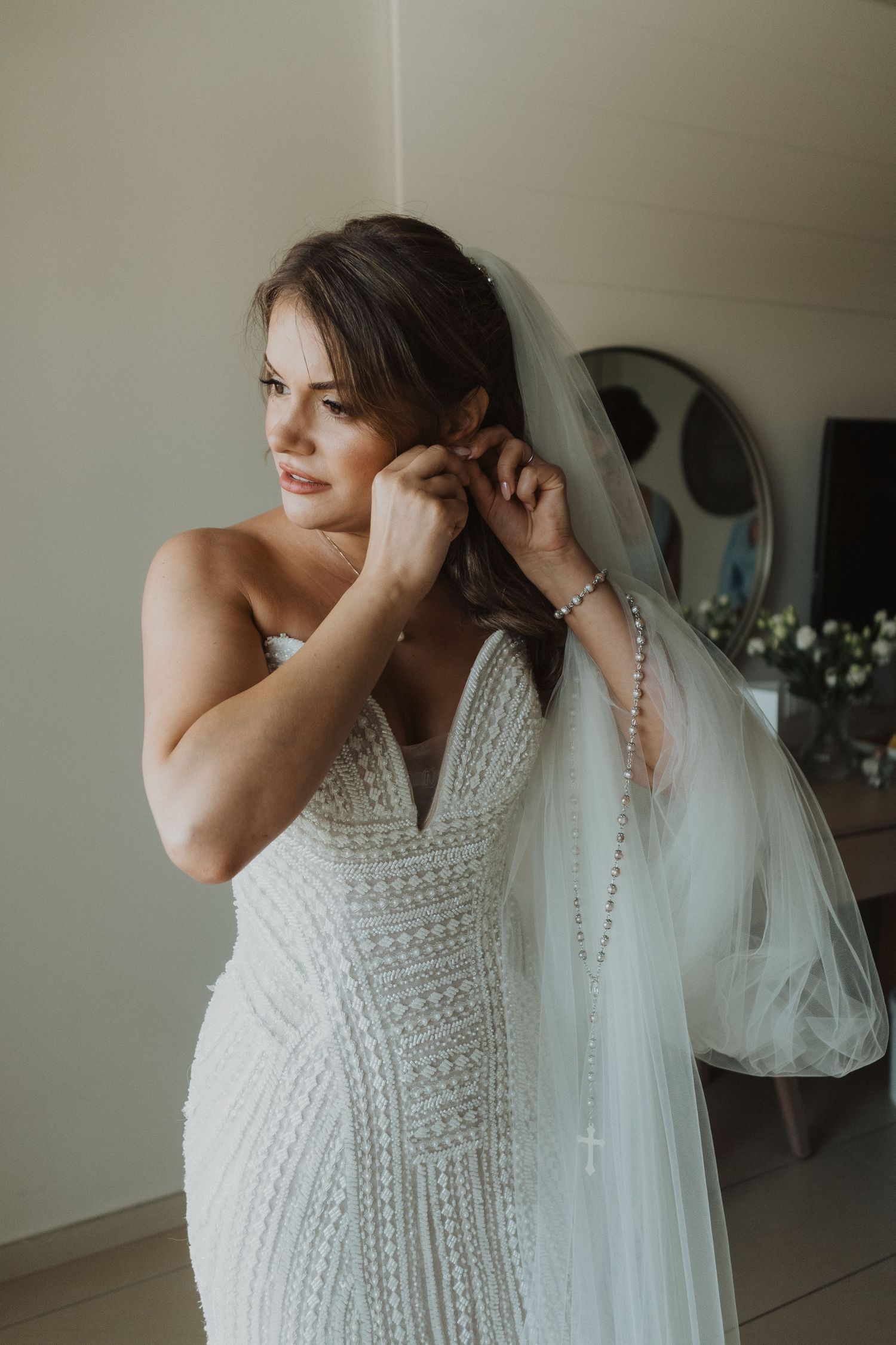 Bride in strapless beaded wedding gown adjusts earrings while wearing veil during bridal preparations.