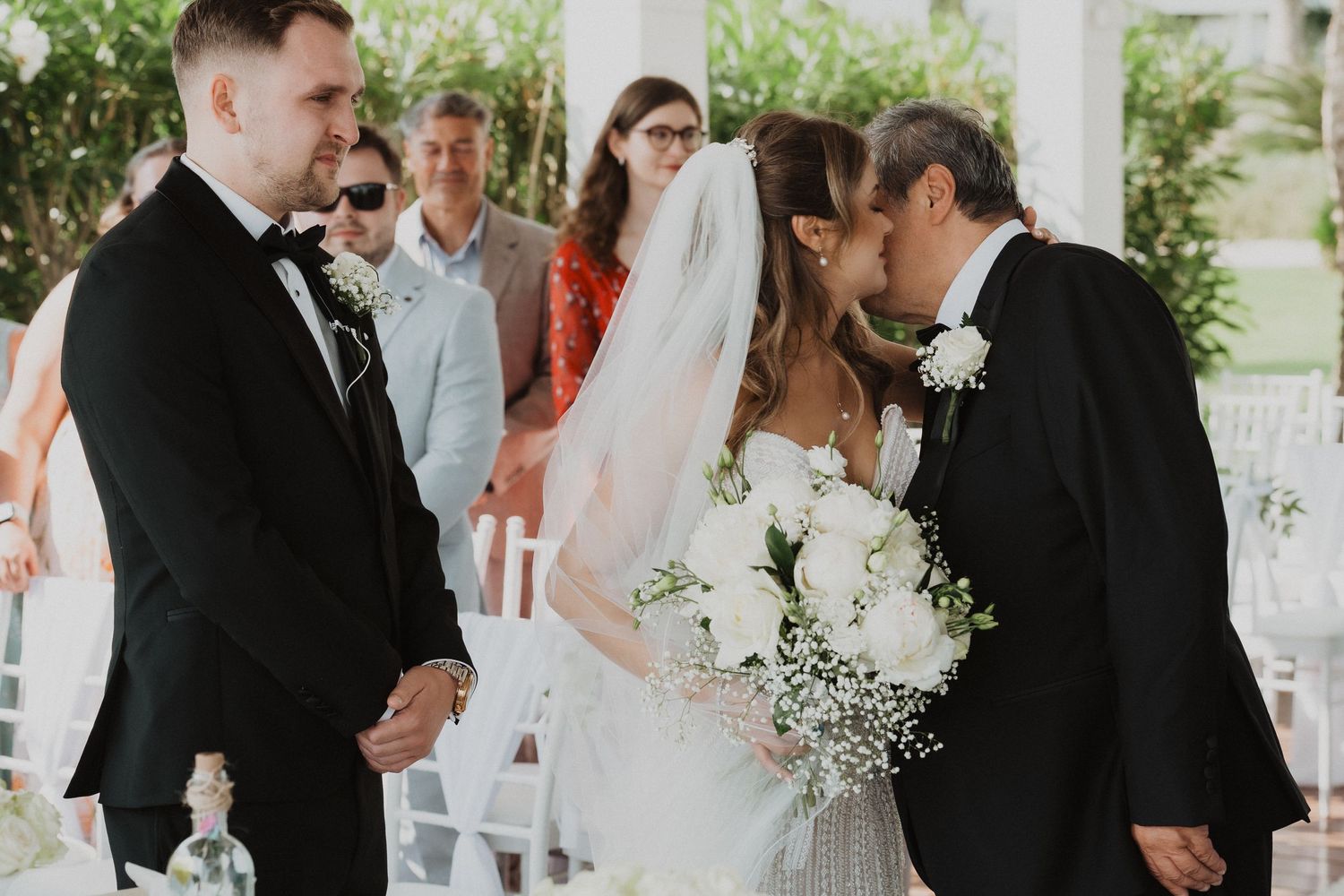 Father of the bride kisses daughter as groom waits at outdoor wedding ceremony altar.