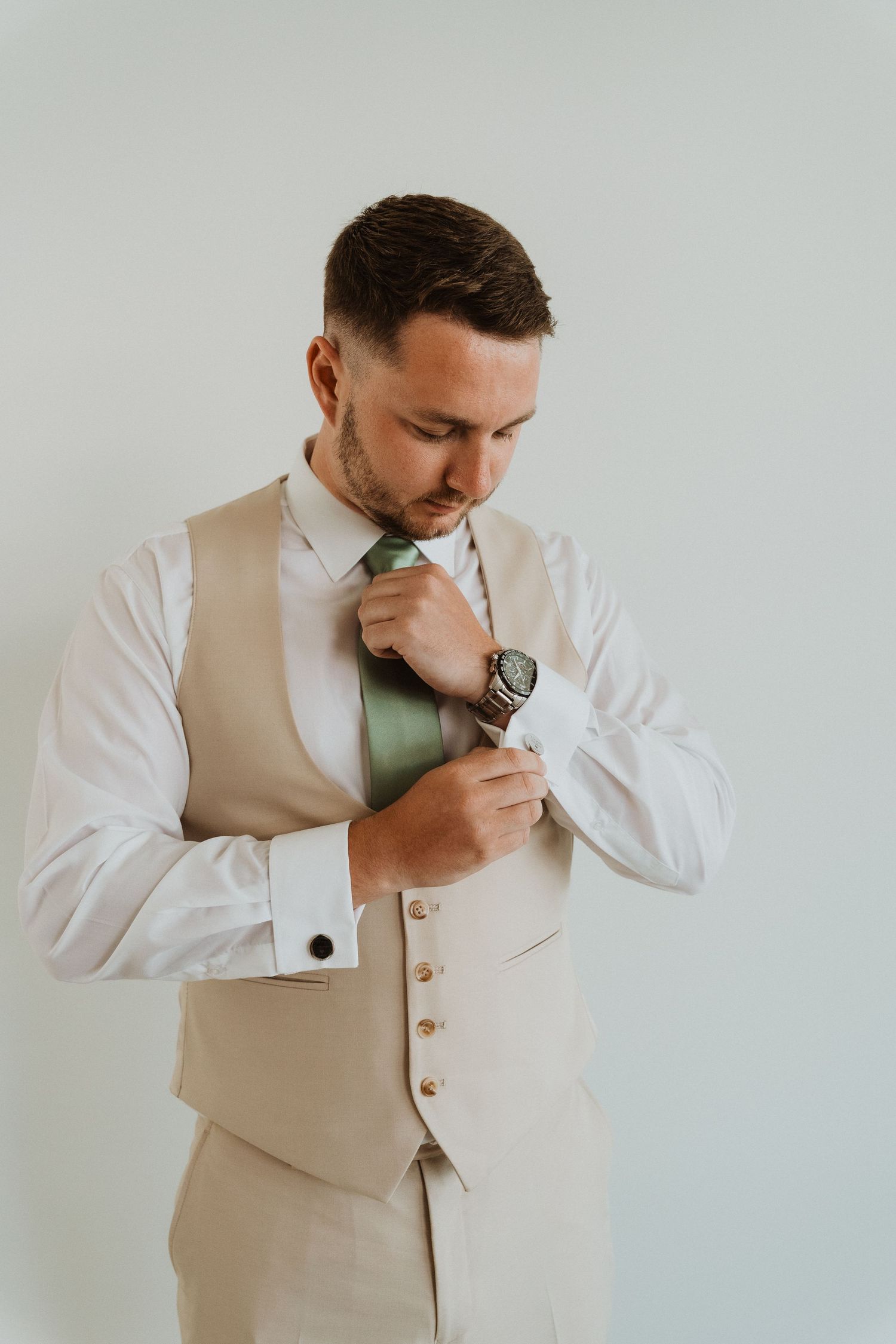 A person in a beige vest and white dress shirt adjusts their green tie while getting dressed.