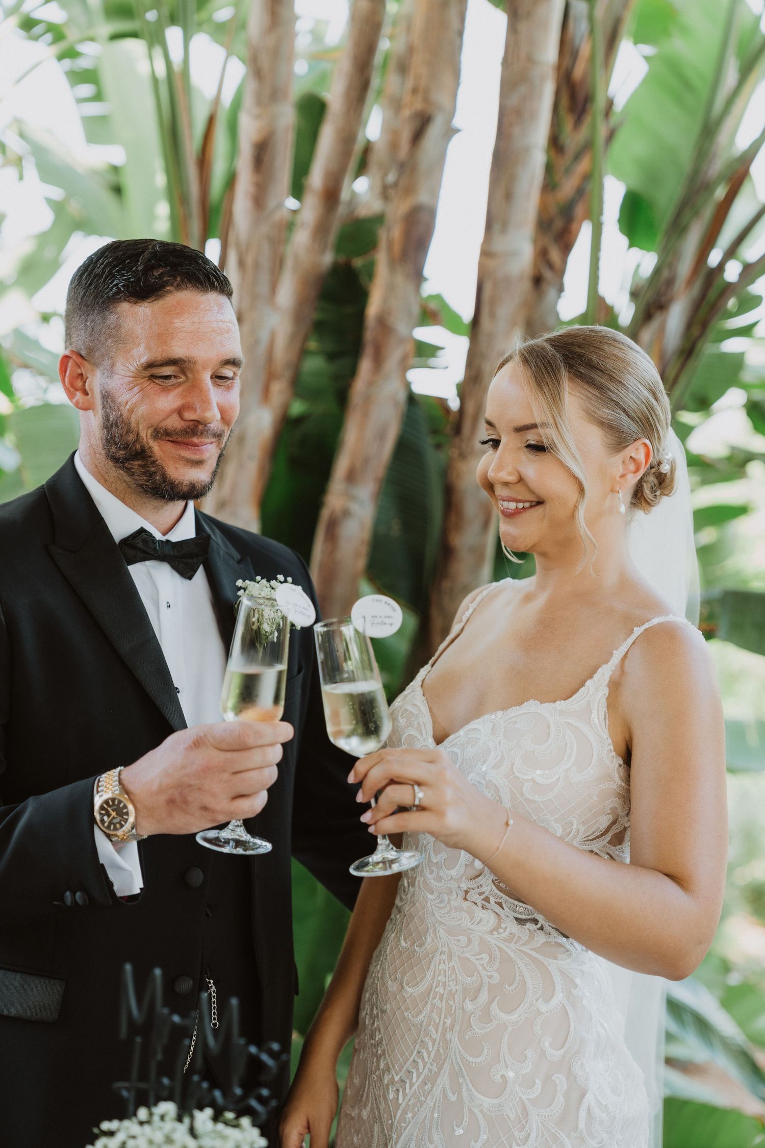 Newlyweds toast with champagne glasses in an outdoor tropical garden setting.