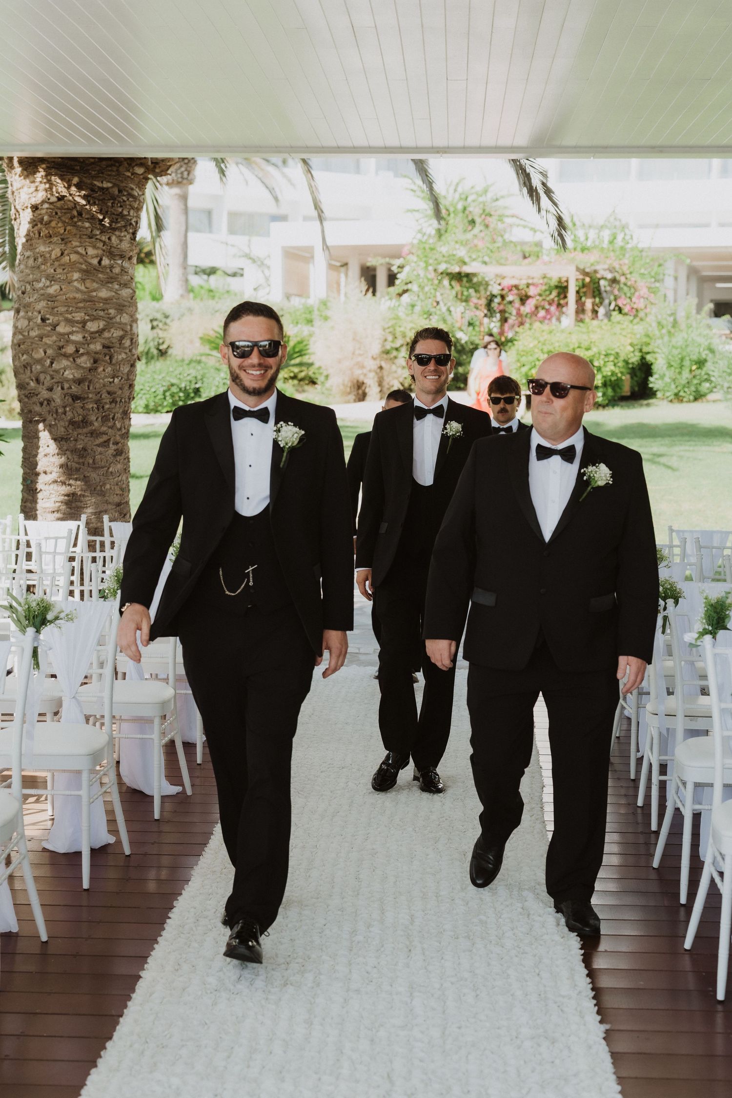 Groomsmen in black tuxedos and sunglasses walk together on a covered outdoor walkway lined with white chairs.