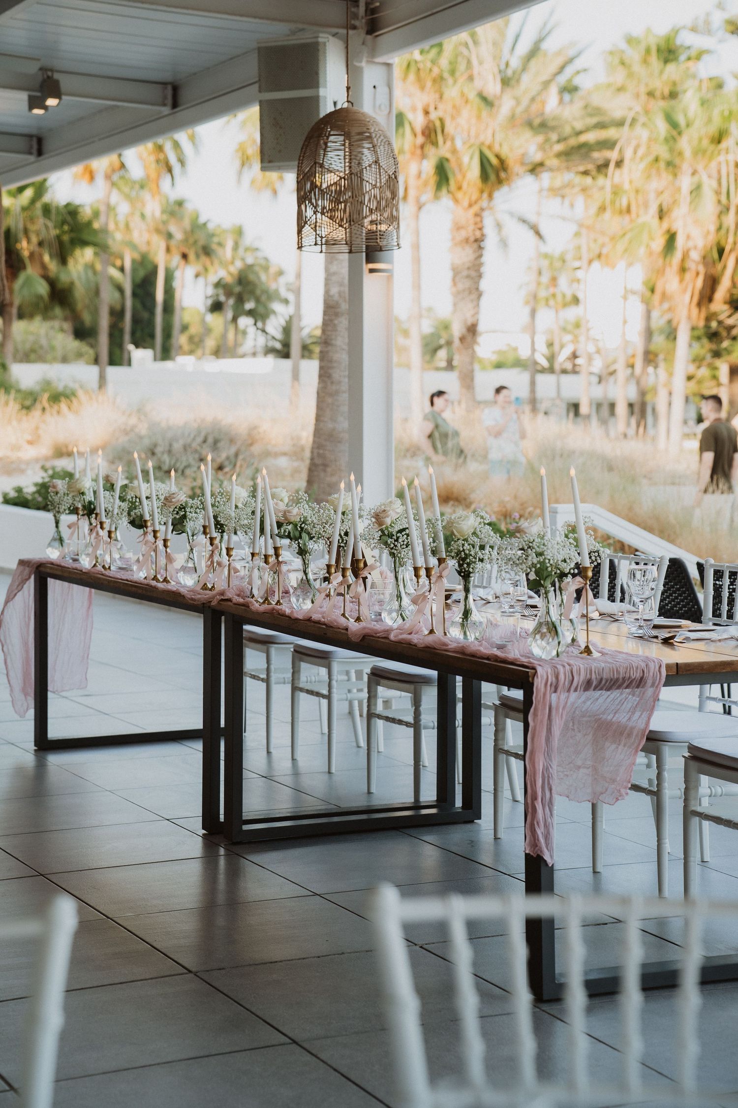 Elegant wedding reception tables with white chairs and floral centerpieces set up in an outdoor covered venue with palm trees.
