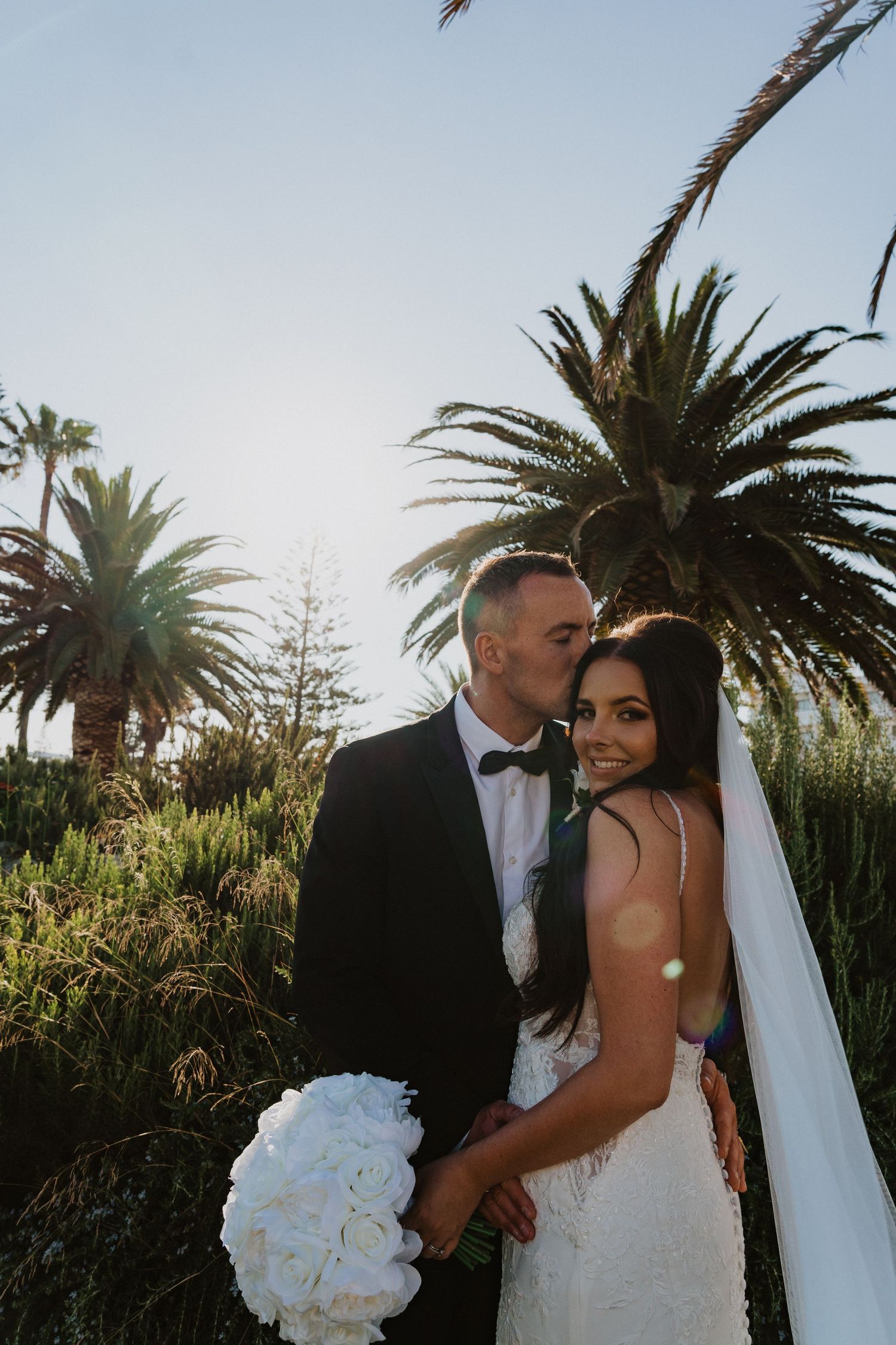 A newlywed couple poses together in a garden with palm trees during golden hour.