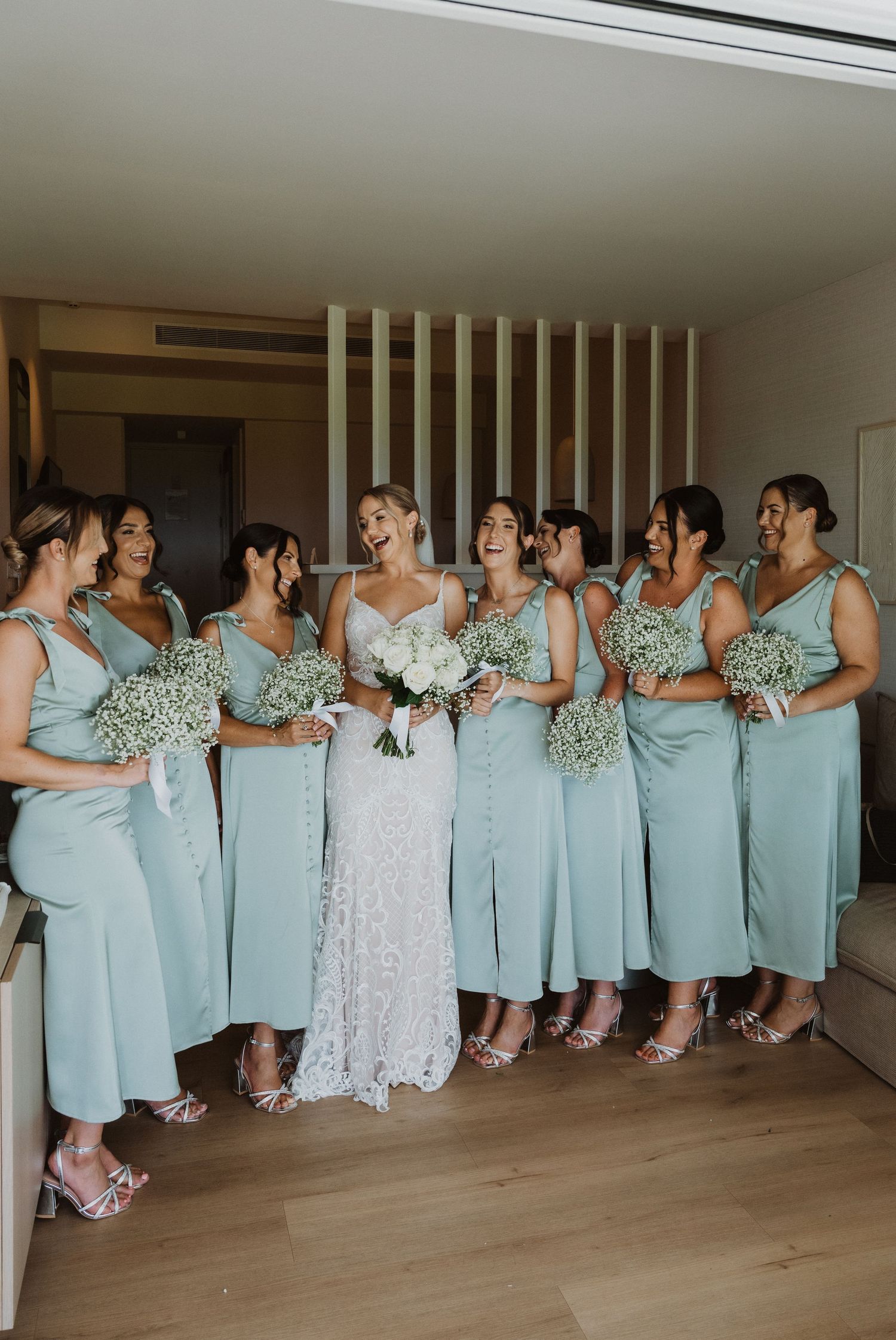 A row of bridesmaids in light blue dresses pose together against white walls in a panoramic wedding photo.