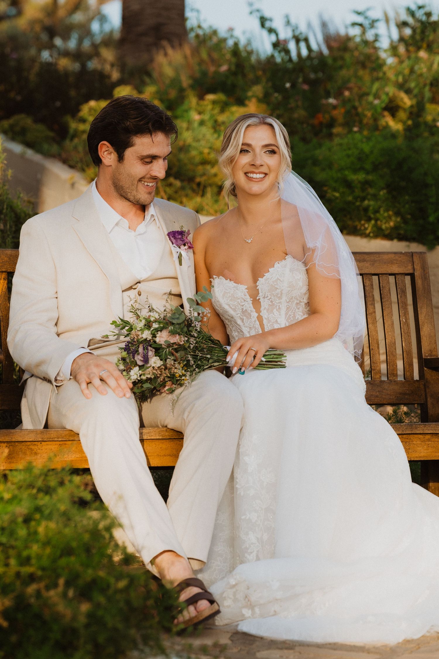 Wedding couple shares joyful moments while sitting on a wooden bench in a garden setting.