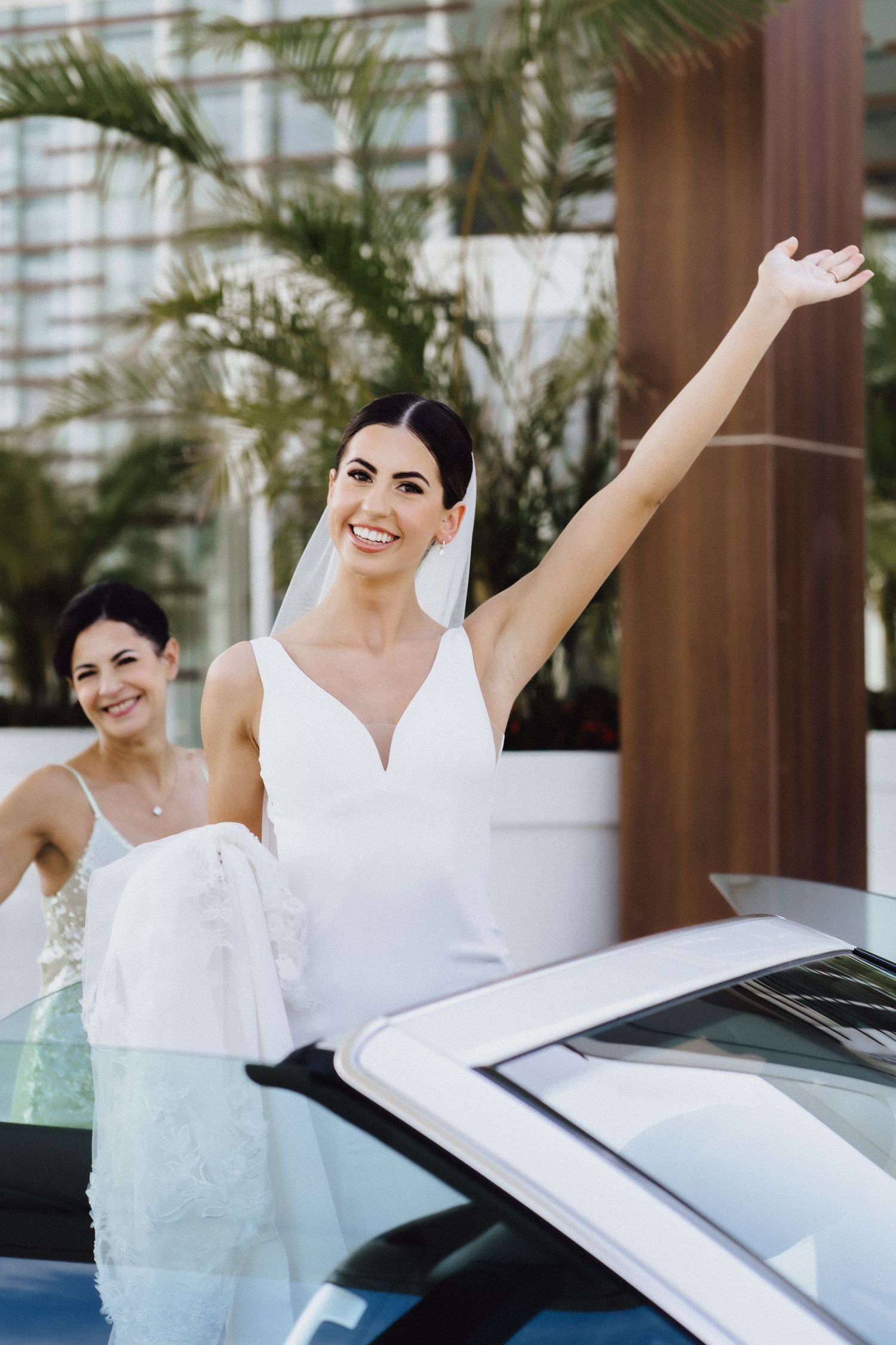 Joyful wedding moment captured near luxury car with palm trees in background.