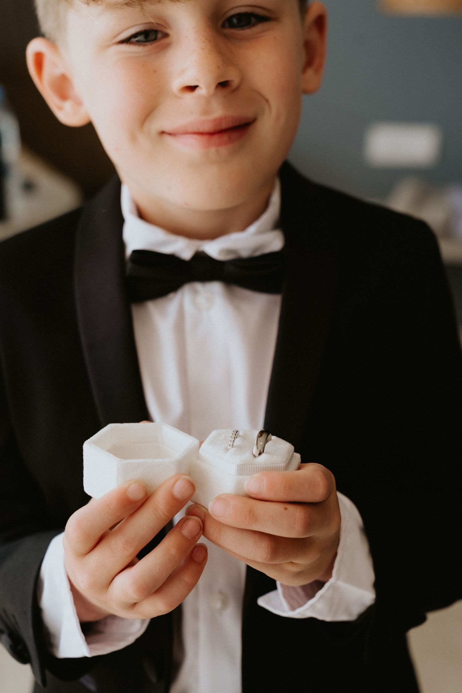 A young ring bearer in formal attire holds a white ring box.