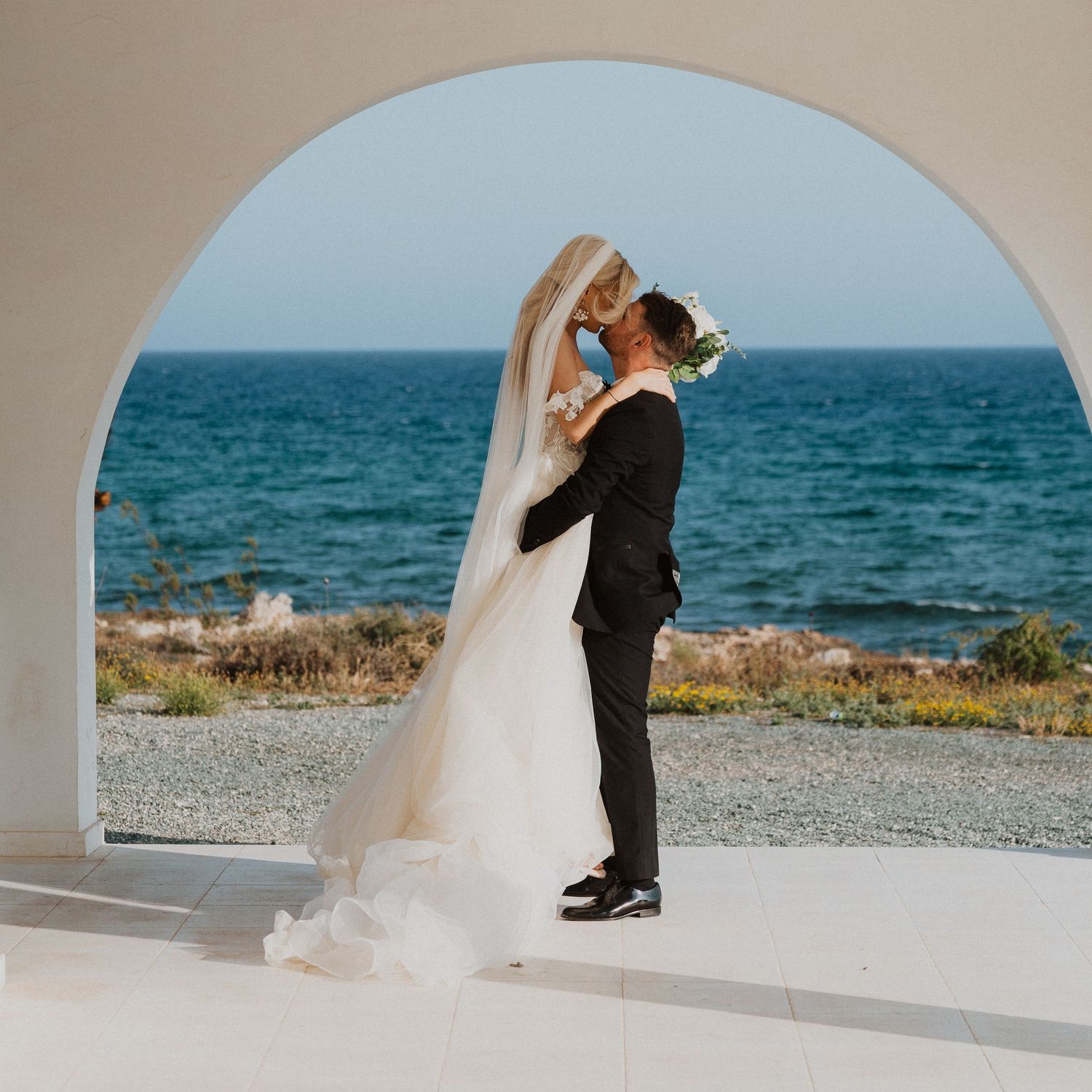 A romantic wedding kiss framed by a white arch with the Mediterranean Sea in background.