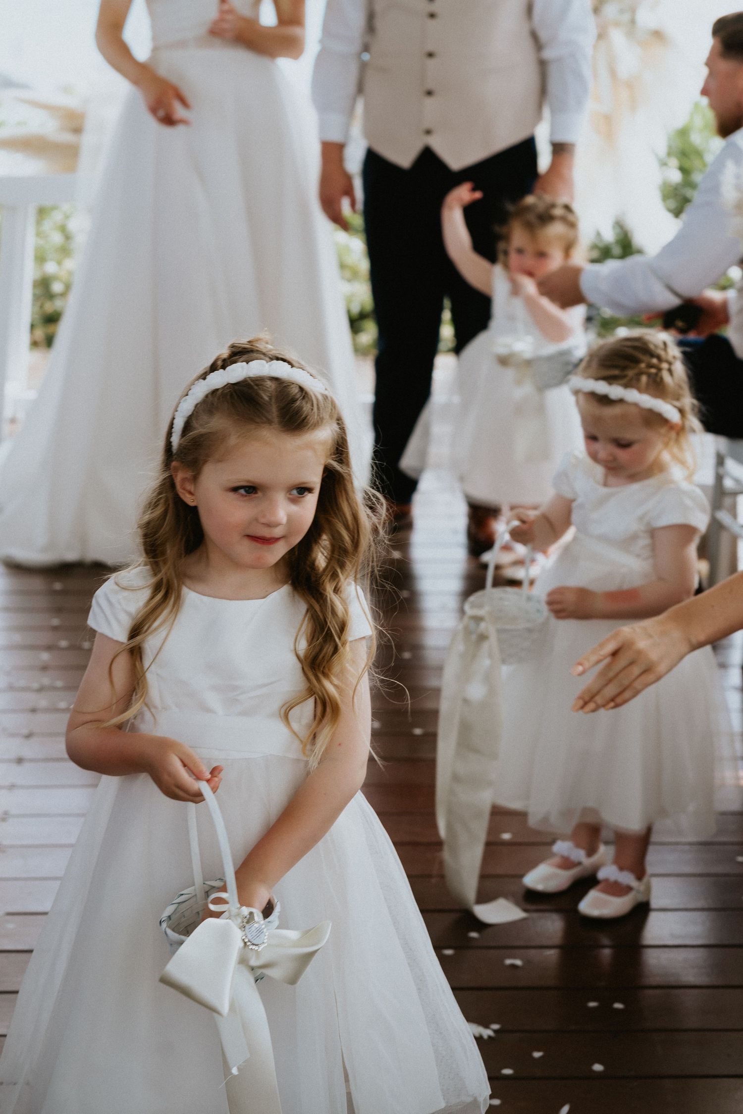 Young flower girls in white dresses carry baskets during waterfront wedding ceremony.
