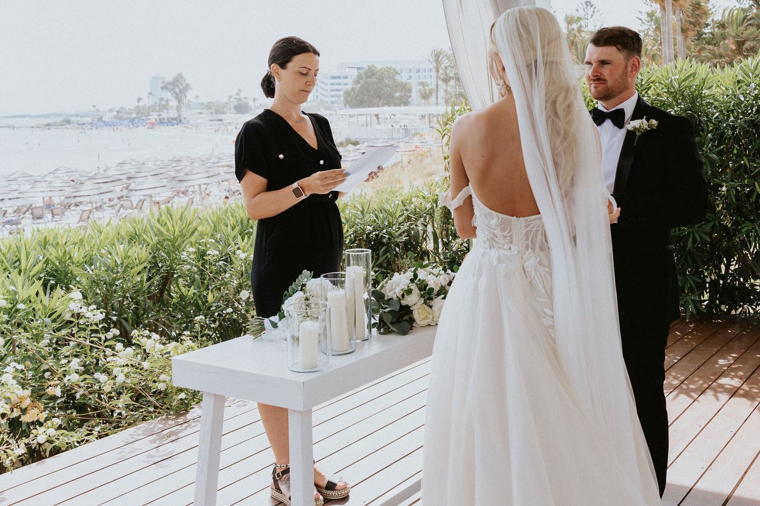 A wedding ceremony takes place on an oceanfront deck with an officiant leading the service.