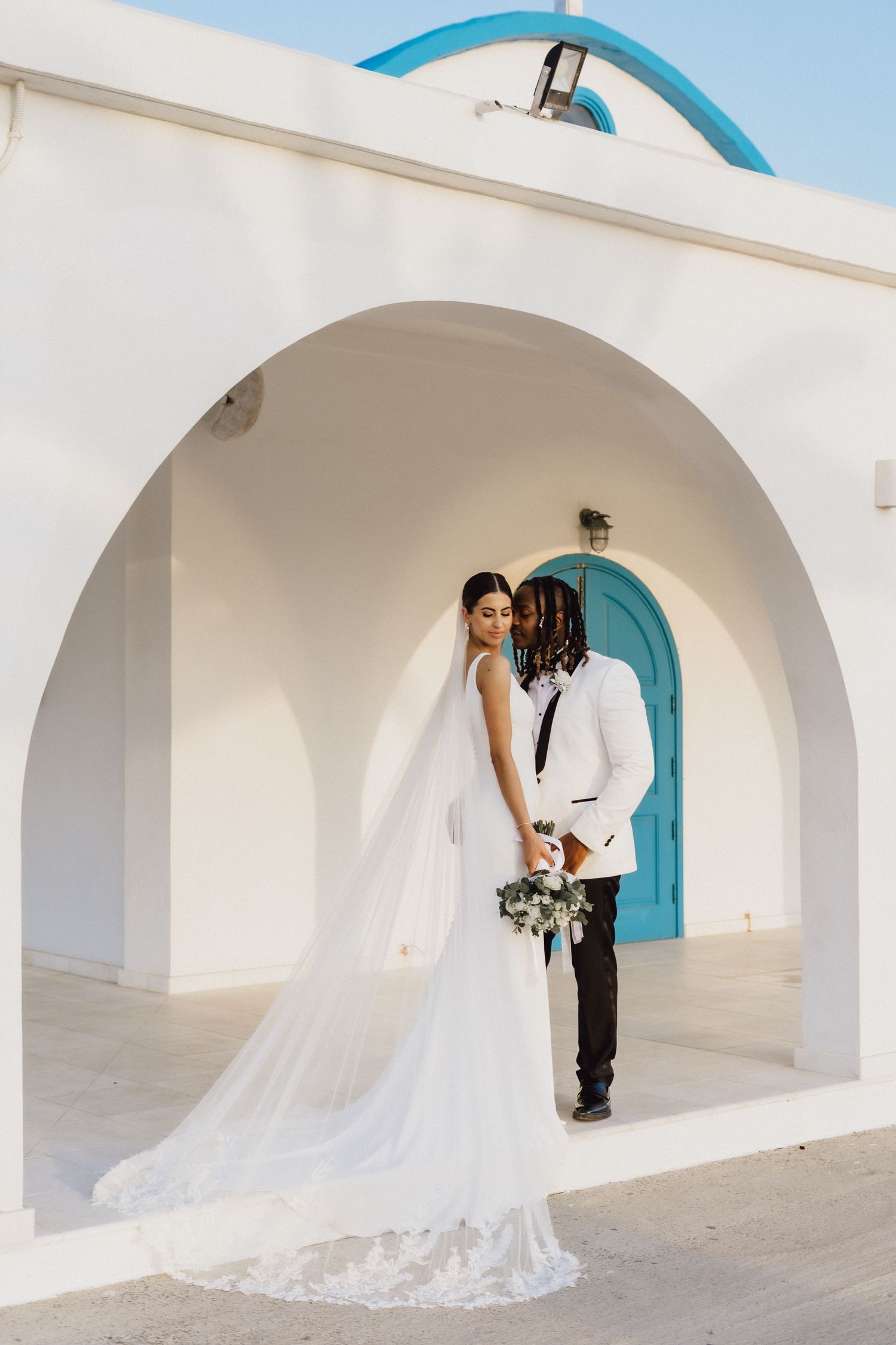 A elegant wedding photo featuring a long flowing veil against white Mediterranean architecture.