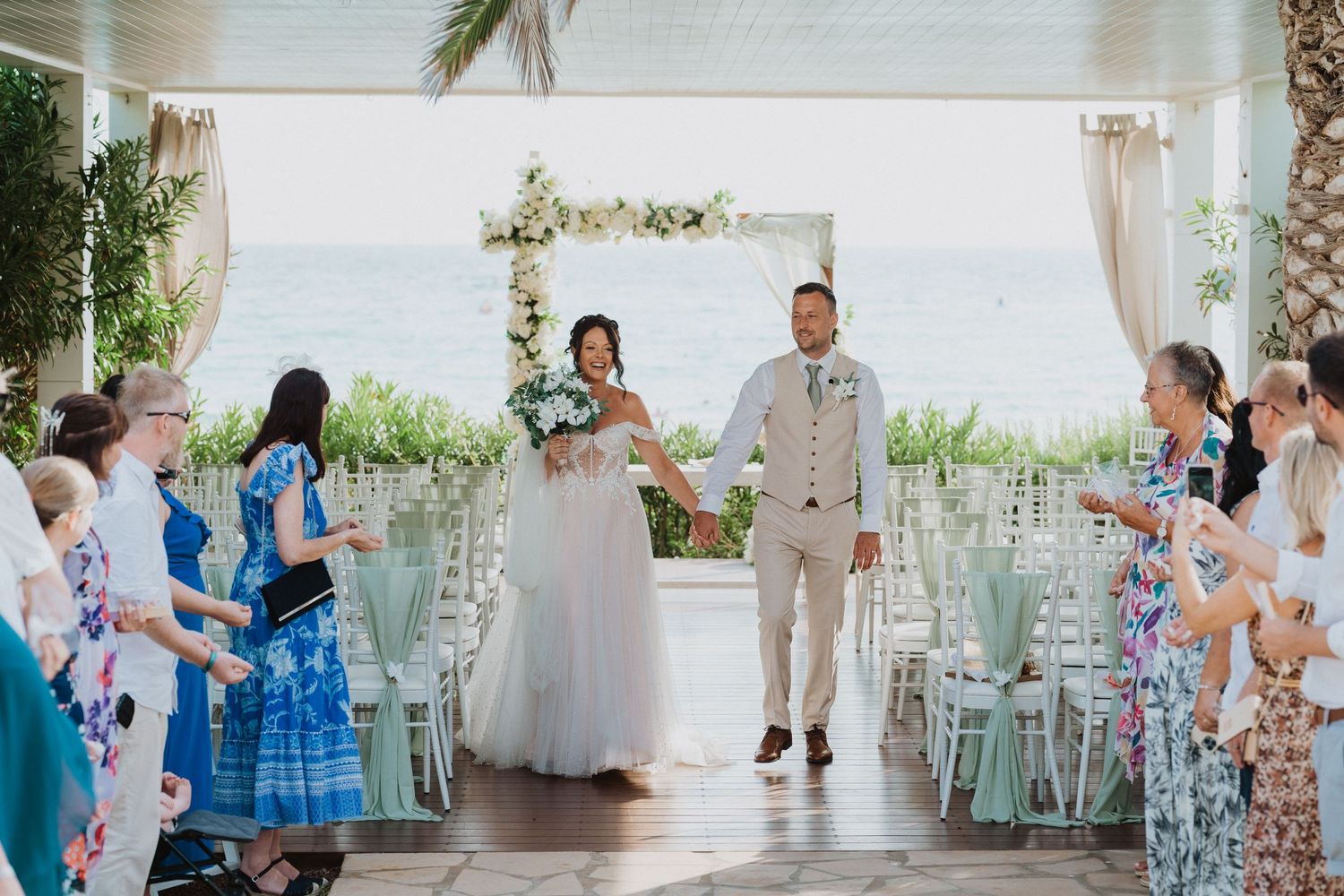 Wedding ceremony at a tropical venue with guests flanking the aisle and turquoise decor chairs.