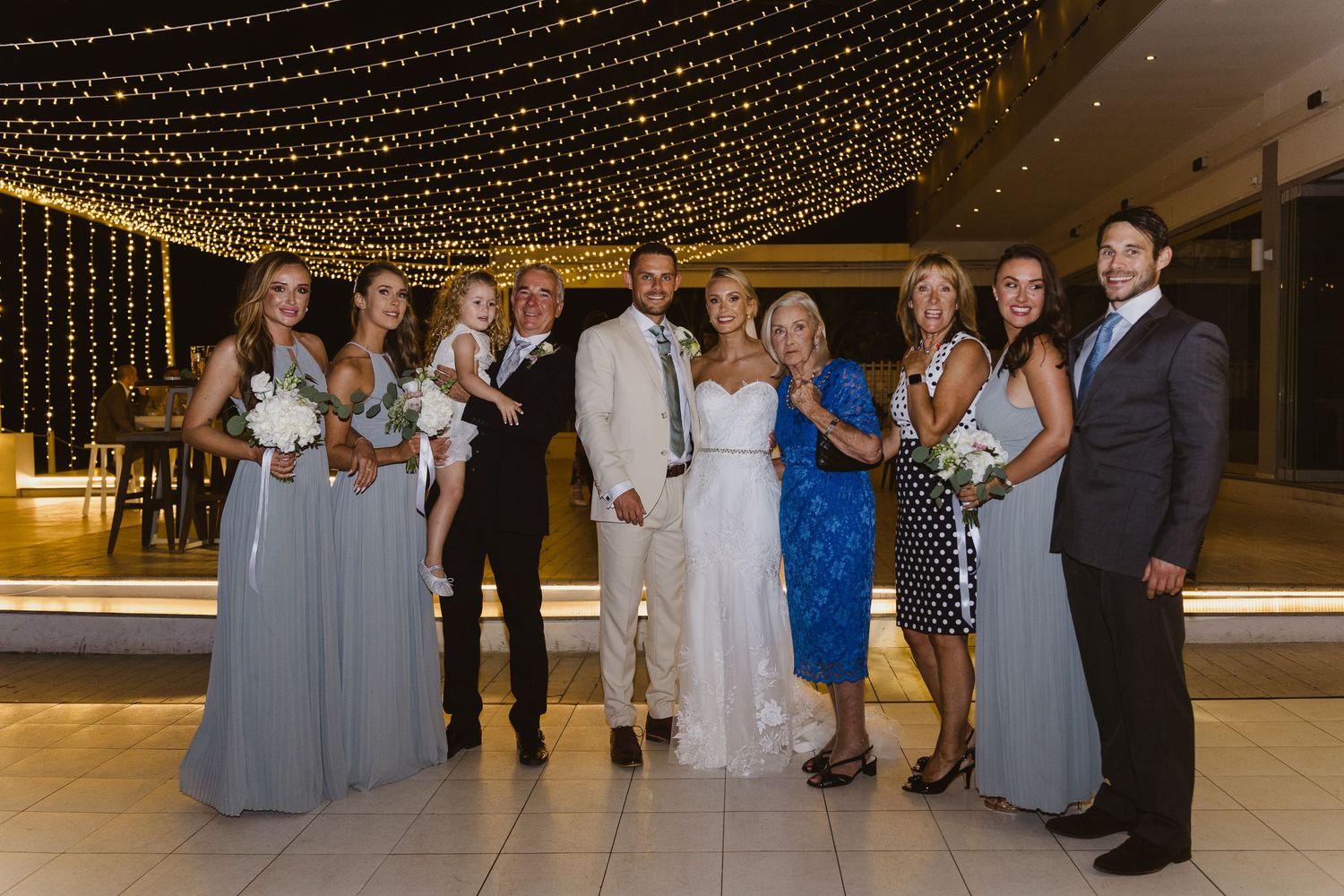 Wedding party poses together under decorative string lights in an elegant venue.