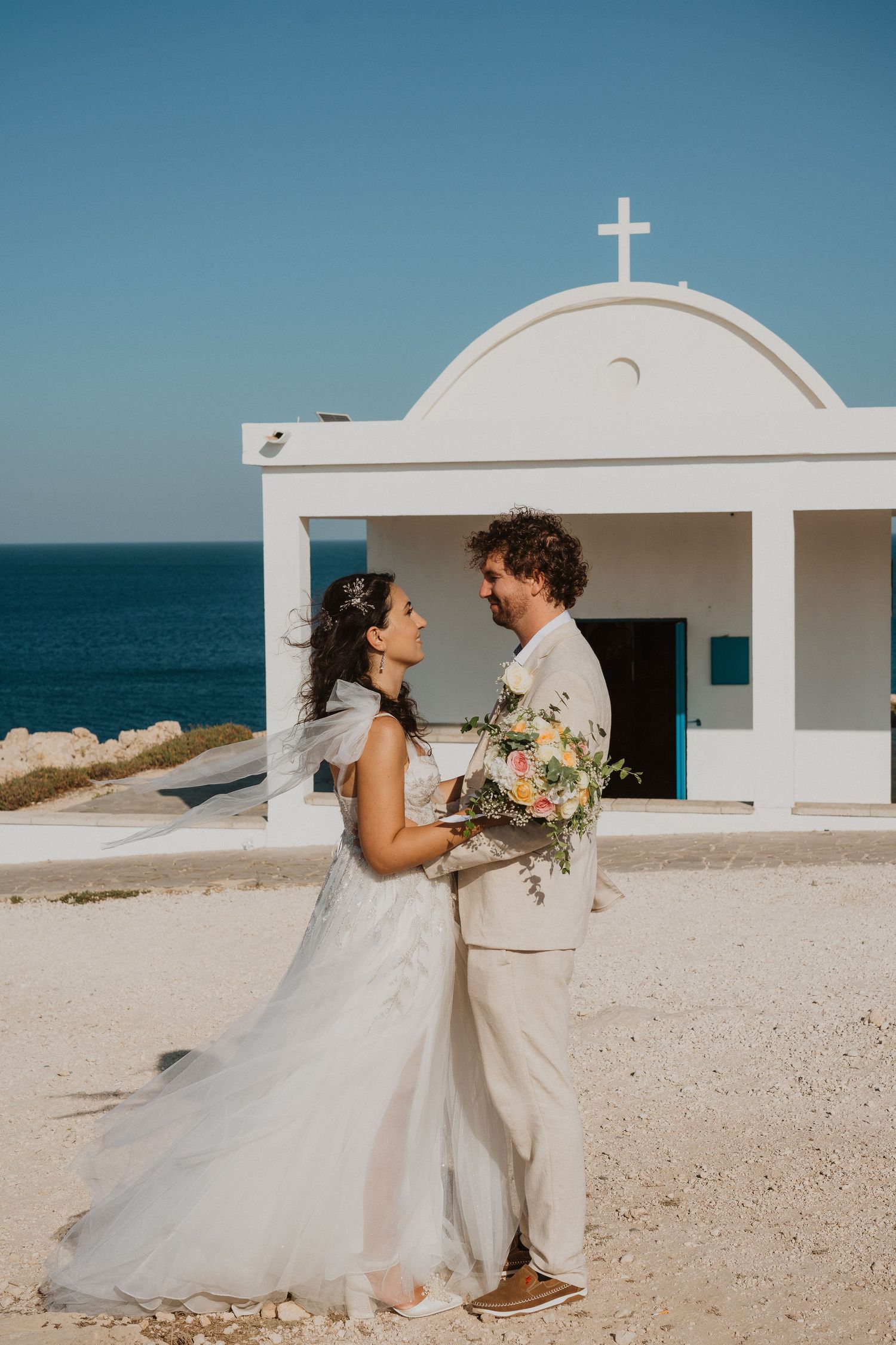A romantic wedding couple embraces in front of a white Mediterranean chapel overlooking the sea.