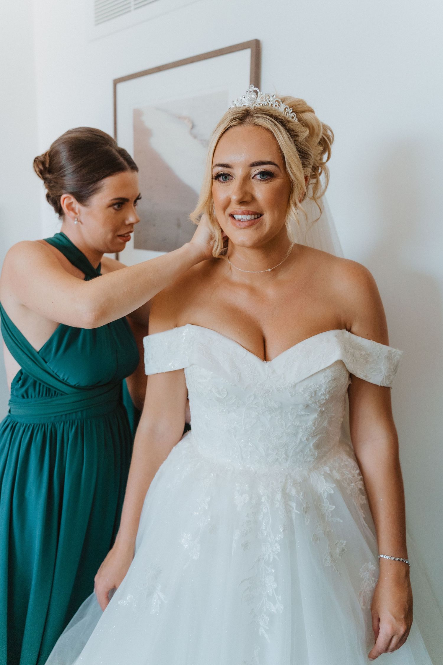 A bride in an off-shoulder white lace gown stands while an attendant in emerald green helps with final preparations.