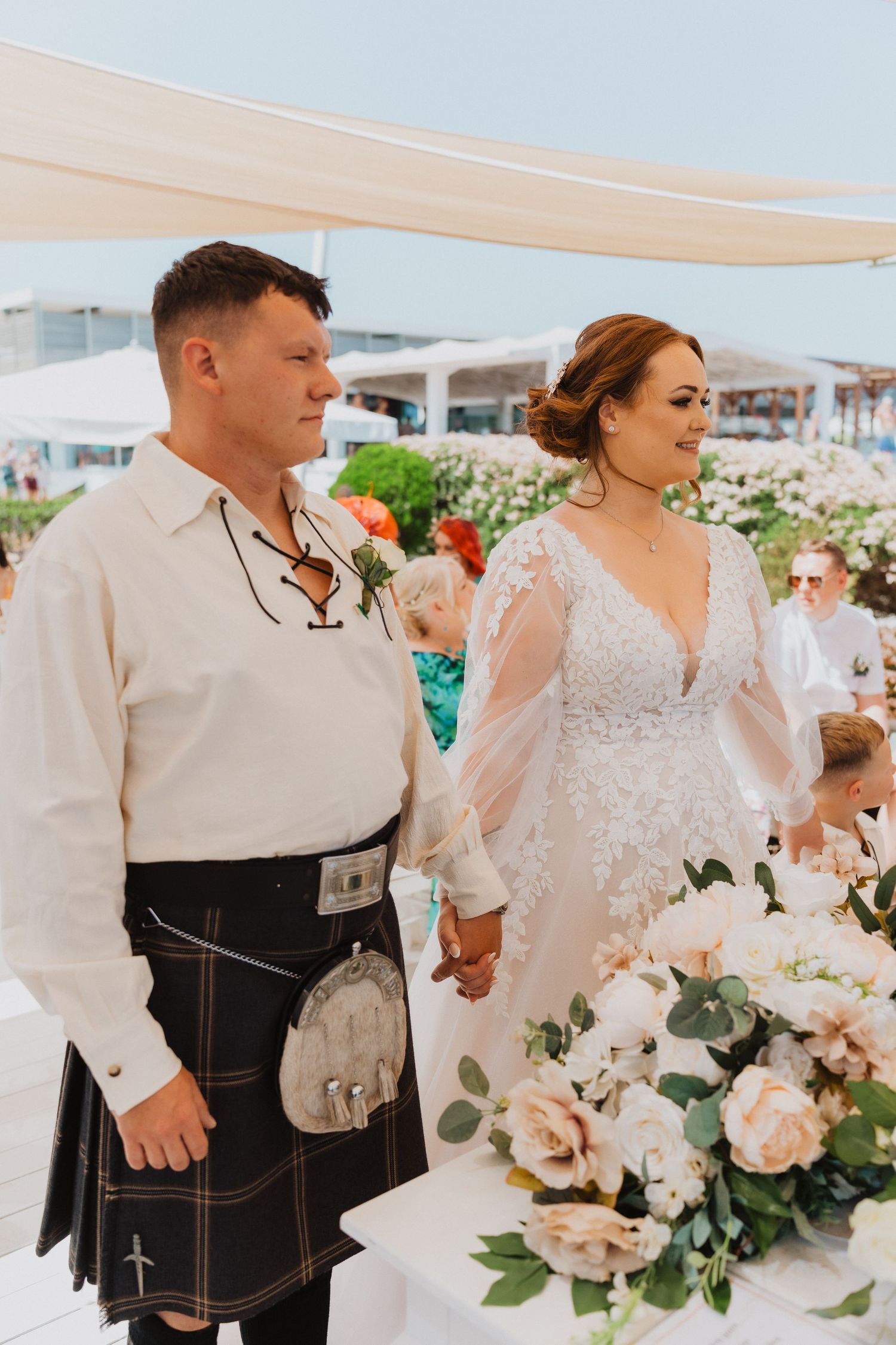 Couple in Scottish and bridal attire standing together at outdoor wedding reception.