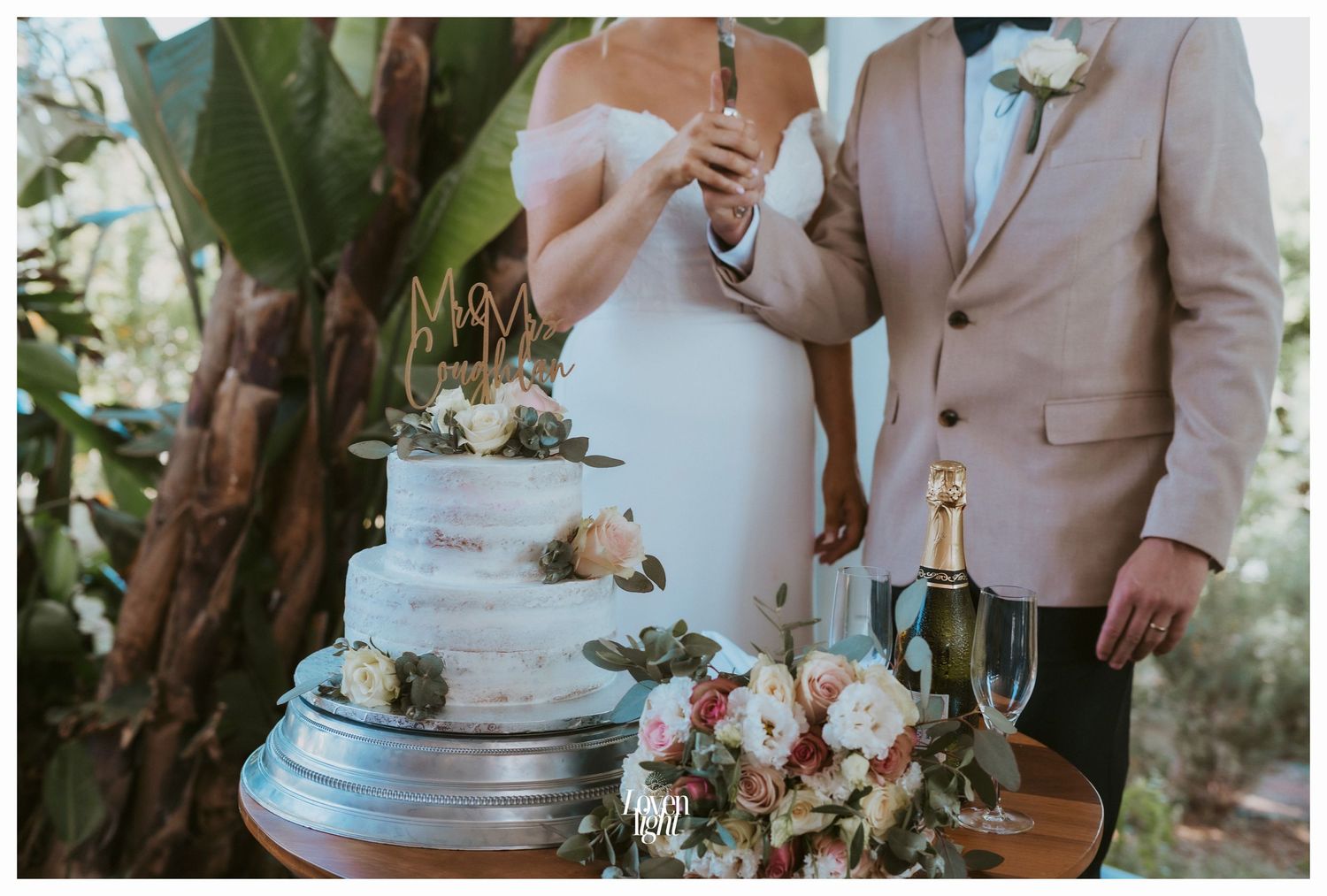 Wedding couple cutting a tiered white cake decorated with flowers on a round wooden table.