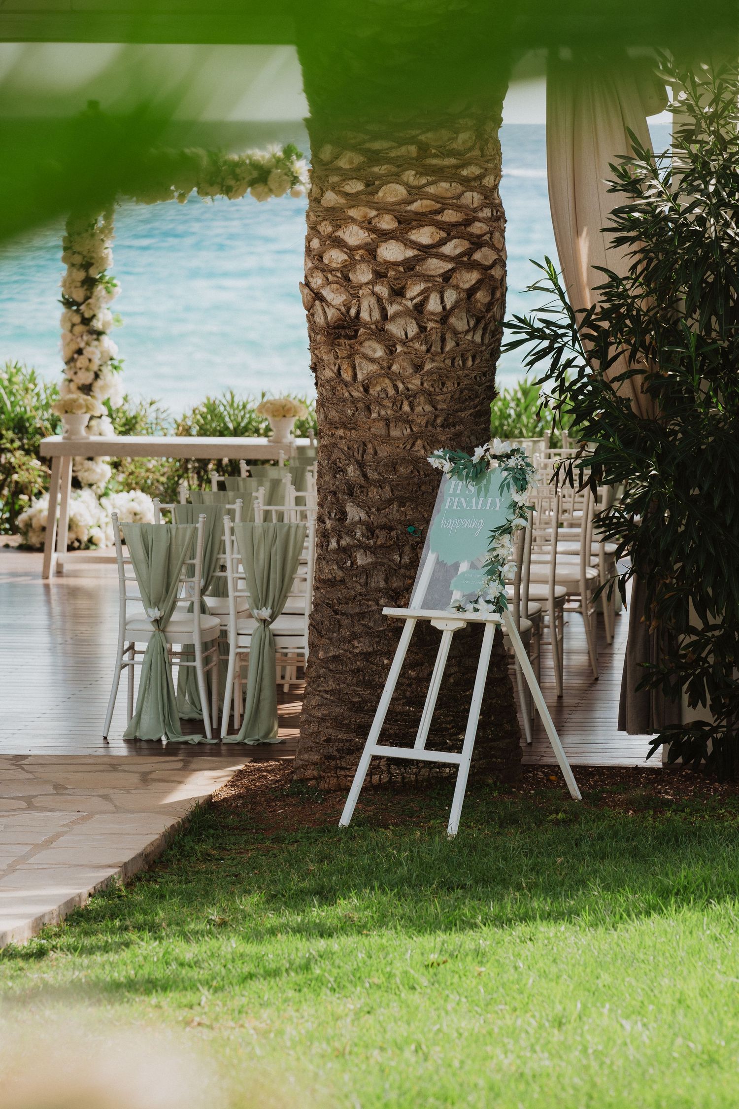 Elegant outdoor wedding setup with palm trees, glass vases on stands, and a glimpse of ocean views through a patio.