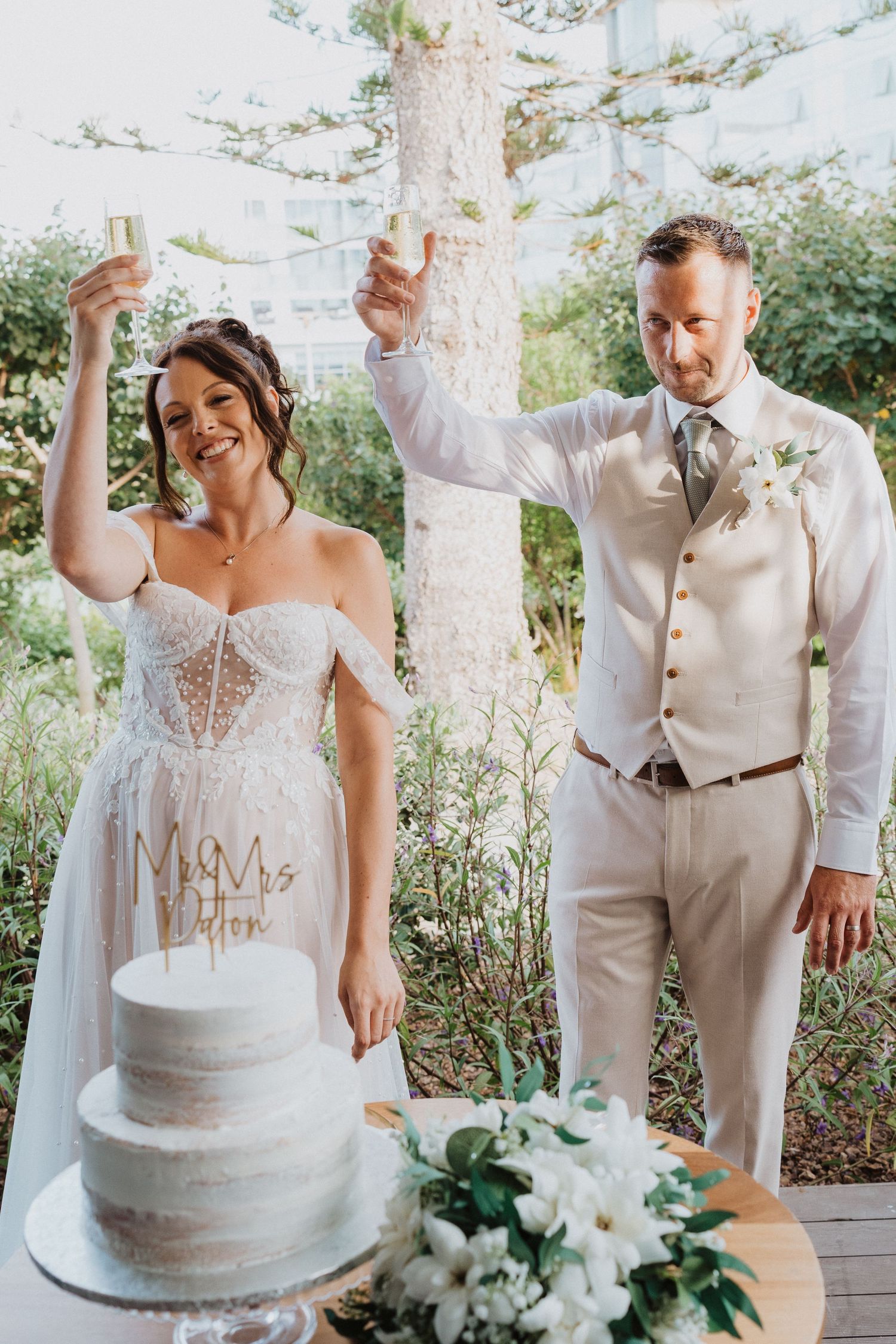 A couple celebrates joyfully at their outdoor garden wedding reception next to their elegant white wedding cake.
